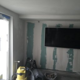 MajsterReda - Pokój w trakcie prac wykończeniowych: ściany z nałożoną gładzią, widoczny telewizor, drabina, odkurzacz i legowisko dla psa. Etap przygotowania do malowania.