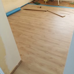 MajsterReda - Układanie paneli podłogowych w jasnym odcieniu drewna, widoczne narzędzia i materiały przygotowane do montażu, niebieska folia izolacyjna przy ścianach.