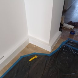 MajsterReda - Białe listwy przypodłogowe i narożnik po malowaniu, zabezpieczone folią malarską. Widoczny nóż do tapet. Podłoga drewniana, w tle płytki.