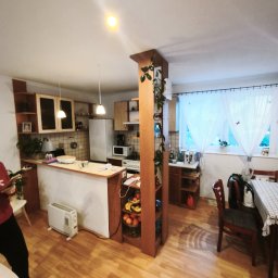 MajsterReda - Jasne panele podłogowe w salonie z aneksem kuchennym w Redzie. Widoczny fragment osoby, stół z krzesłami i kolumna z roślinami.