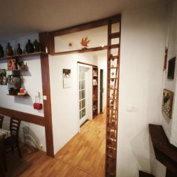 MajsterReda - Jasne wnętrze z drewnianą podłogą i dekoracyjną ścianką działową z drewnianymi elementami. Widoczne półki z dekoracjami i drzwi w głębi.