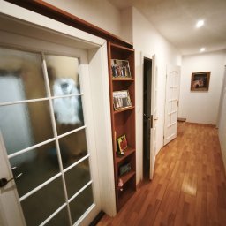 MajsterReda - Korytarz po remoncie: drewniana podłoga, białe drzwi z szybami, regał z dekoracjami. Ciepłe oświetlenie, widok w głąb mieszkania.