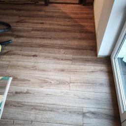 MajsterReda - Ułożone panele podłogowe w odcieniach brązu, widoczne przycięte listwy przypodłogowe, fragment odkurzacza i drzwi. Układanie paneli z widocznymi detalami wykończenia.