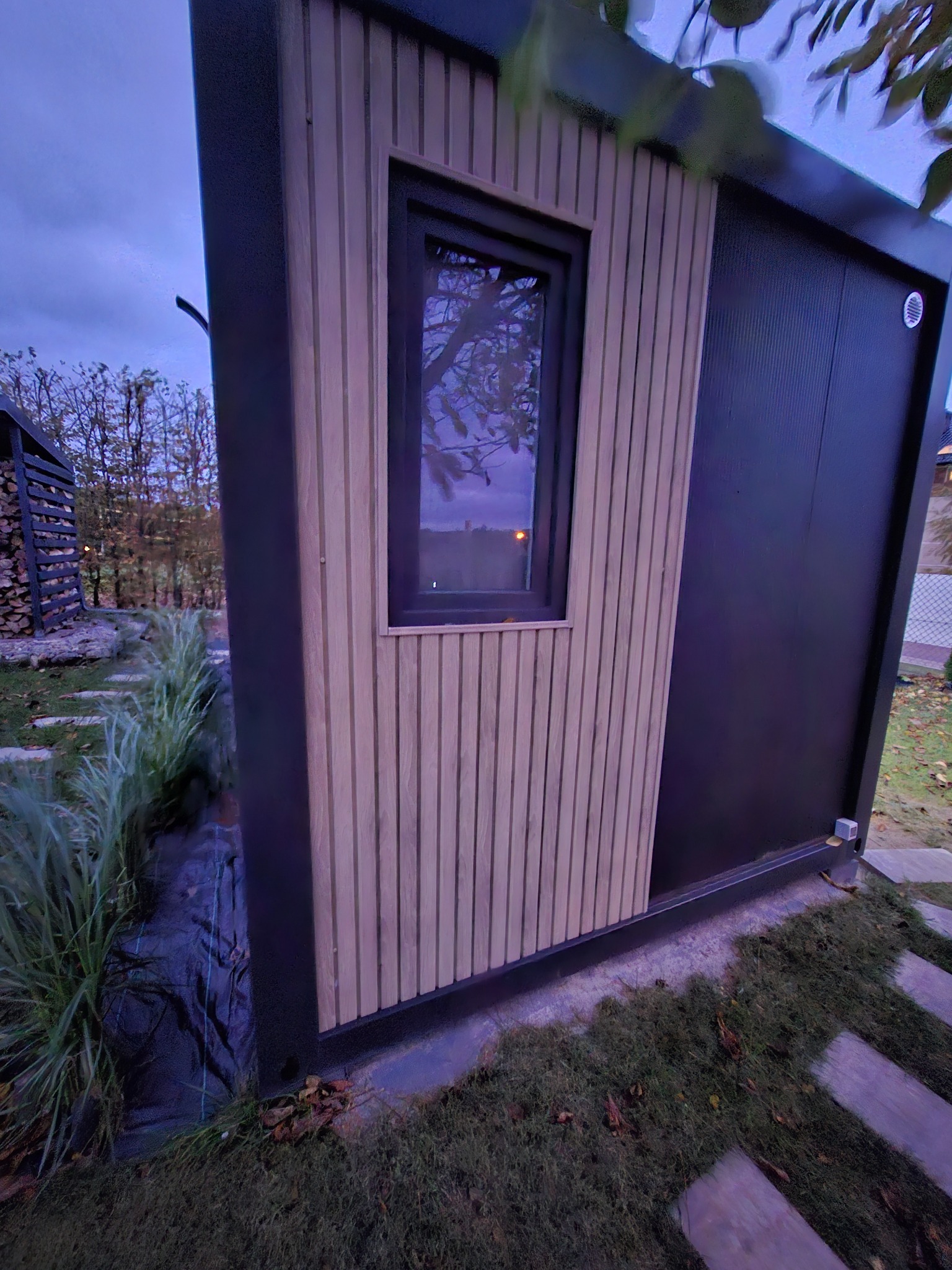 Nowoczesna, modułowa konstrukcja z drewnianą okładziną i czarnymi elementami, otoczona zielenią i brukowaną ścieżką, sfotografowana o zmierzchu.