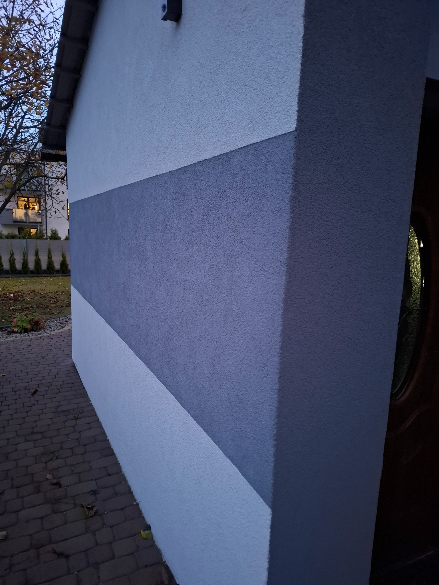 Dwukolorowa elewacja budynku w Redzie z dekoracyjnym pasem, widoczna z perspektywy chodnika z kostki brukowej, oświetlona wieczornym światłem.