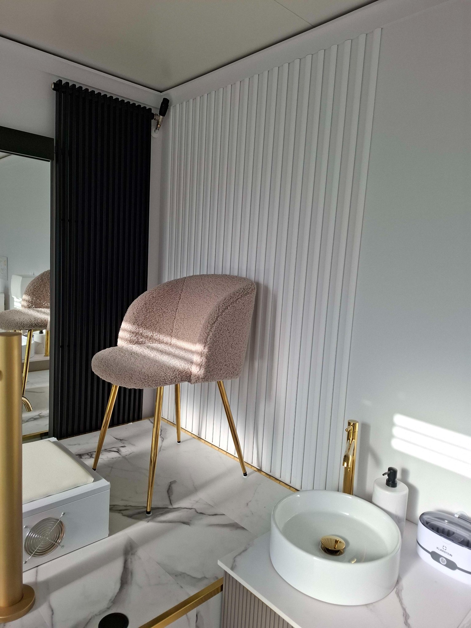 Nowoczesne wnętrze z beżowym krzesłem na złotych nogach, umywalką nablatową z baterią w kolorze złota i dekoracyjnymi panelami ściennymi w odcieniach bieli i czerni.