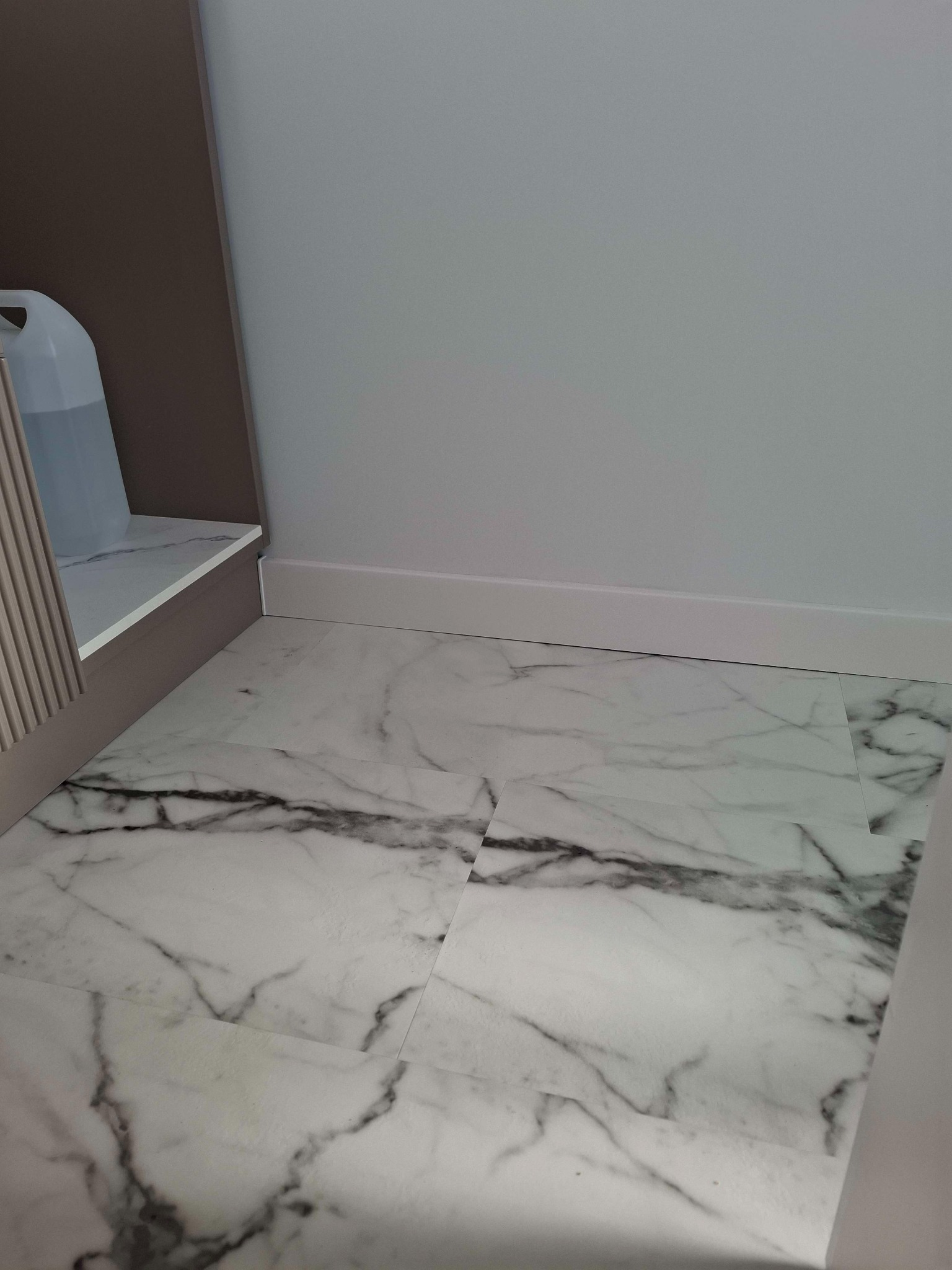 Ułożone białe panele podłogowe imitujące marmur z widocznymi szarymi żyłami, biała listwa przypodłogowa, fragment szafki z beżowymi lamelami i przezroczystym kanistrem.