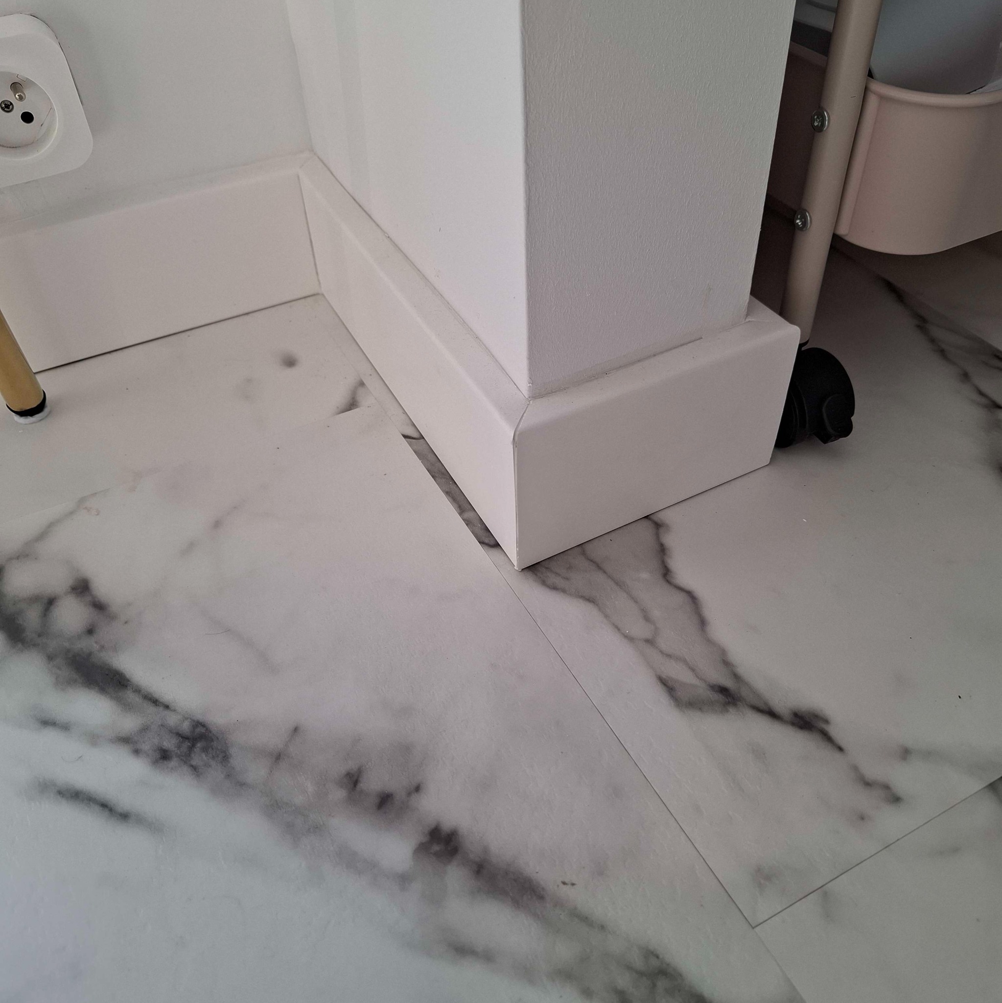 Wnętrze z marmurową podłogą, białą listwą przypodłogową i fragmentem ściany z gniazdkiem elektrycznym oraz kółkiem wózka.