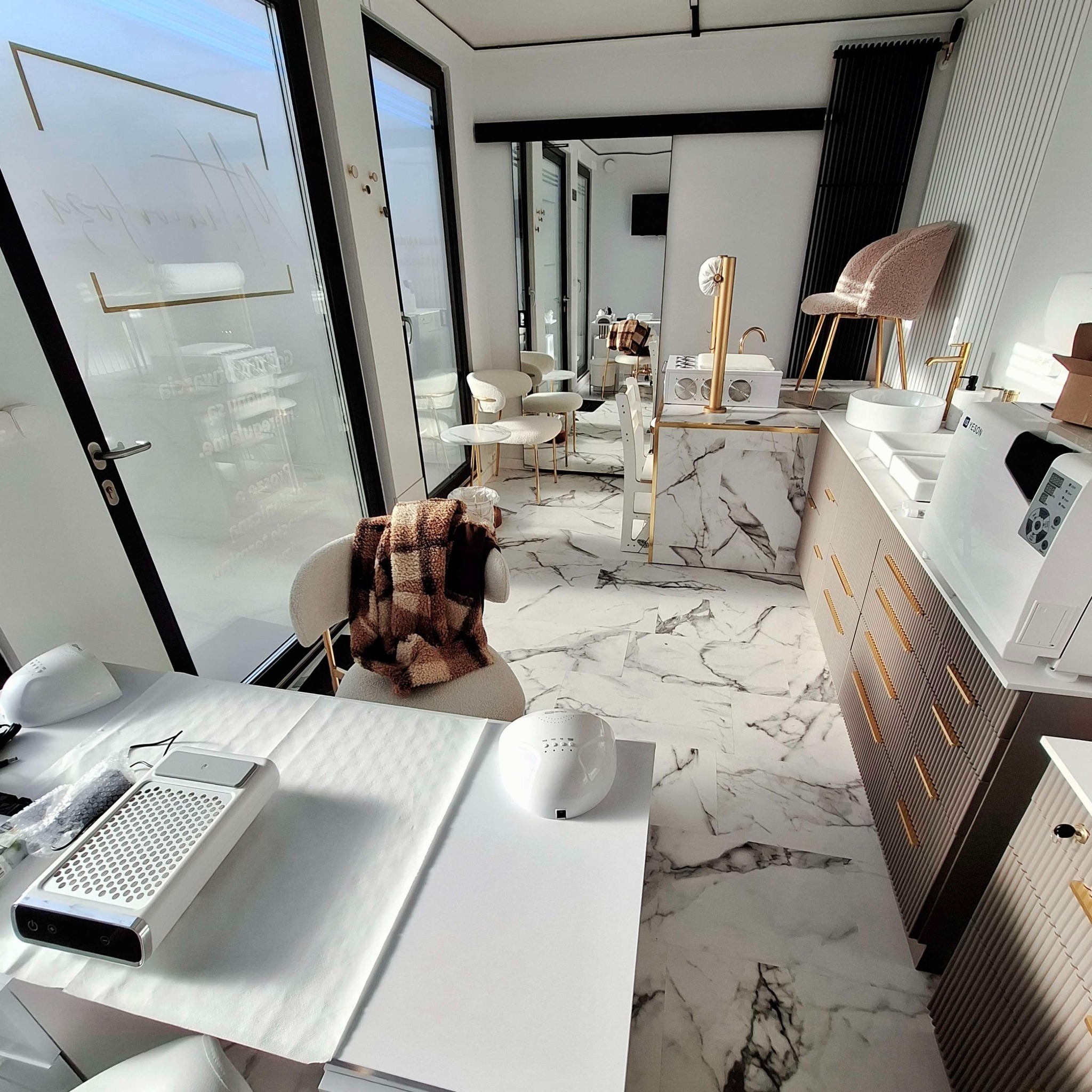 Wnętrze salonu kosmetycznego z marmurową podłogą, stanowiskami do manicure z pochłaniaczami pyłu i lampami UV, oraz krzesłami z pluszową narzutą w kratę.