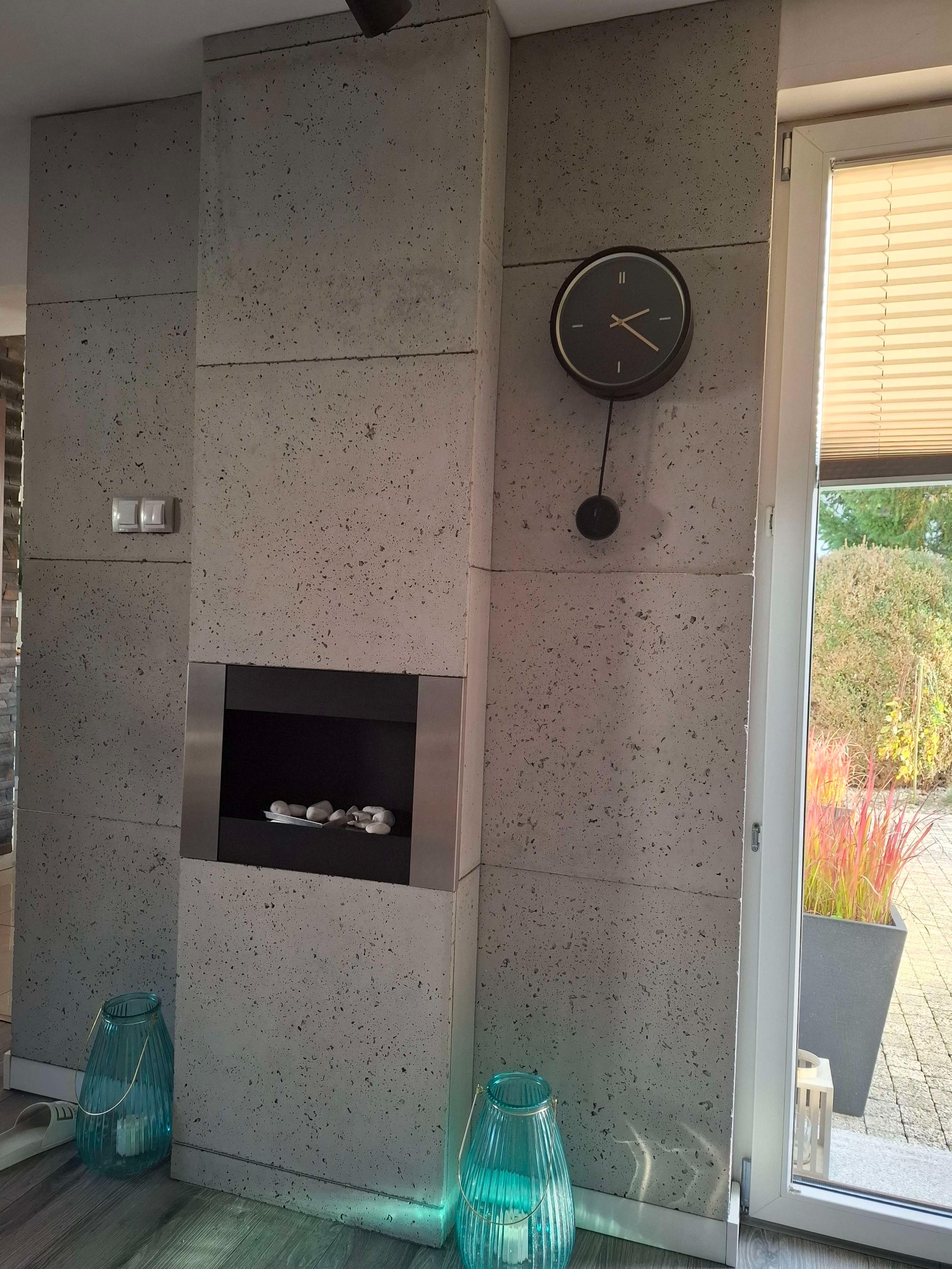 Ściana wyłożona betonowymi płytami z wbudowanym biokominkiem i wiszącym zegarem, dekoracyjne lampiony na podłodze, widok na ogród przez okno.