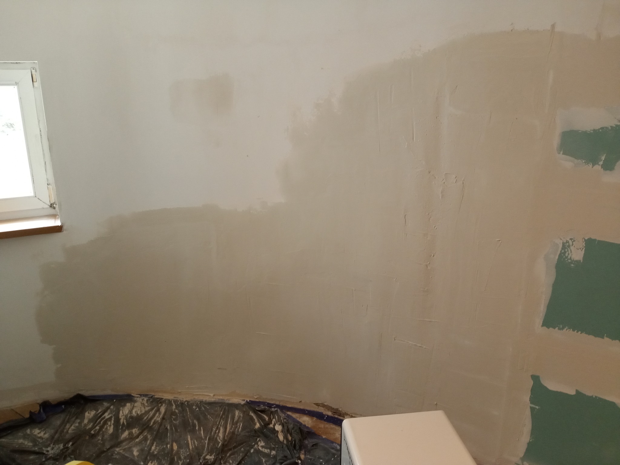 Fragment ściany w trakcie szpachlowania, z widocznym oknem po lewej stronie i zabezpieczoną folią podłogą, ukazujący nierównomierne nałożenie gładzi.