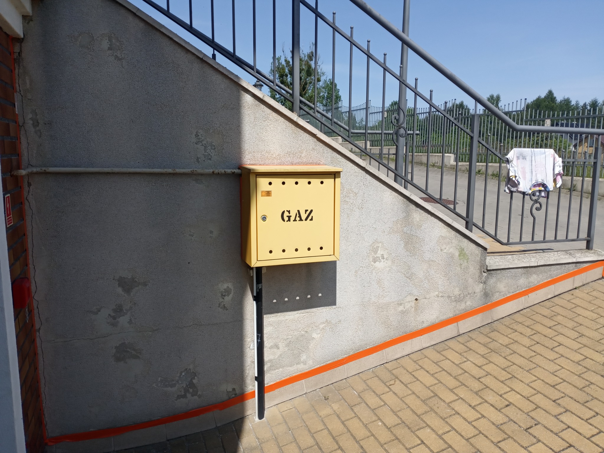 Żółta skrzynka gazowa na szarej, chropowatej ścianie pod schodami z metalową barierką, pomalowana pomarańczową farbą. Widoczne ślady wilgoci na ścianie.