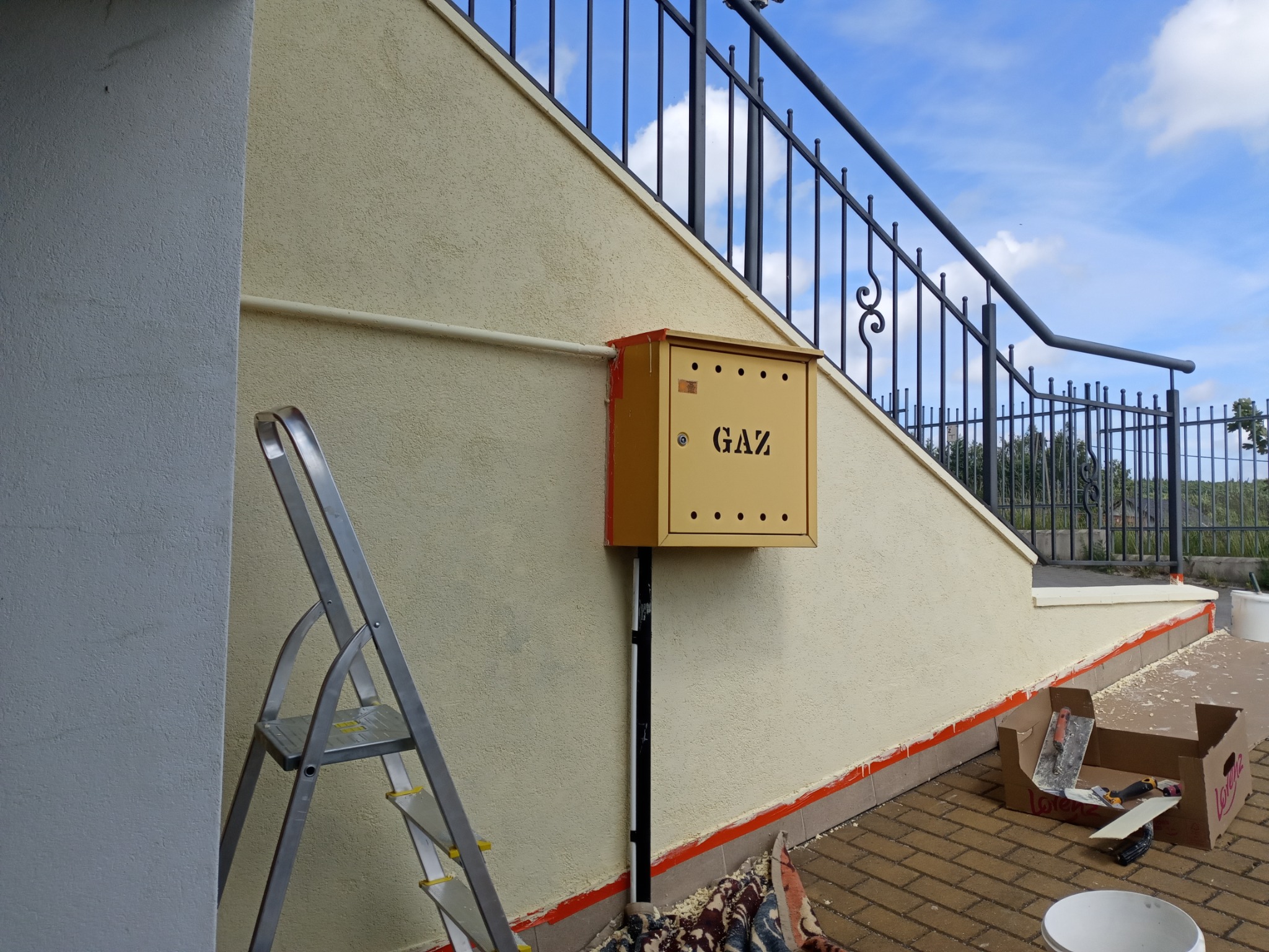 Ściana budynku po malowaniu z żółtą skrzynką gazową, widoczne narzędzia malarskie i drabina. Świeżo pomalowana elewacja w ciepłym odcieniu beżu.