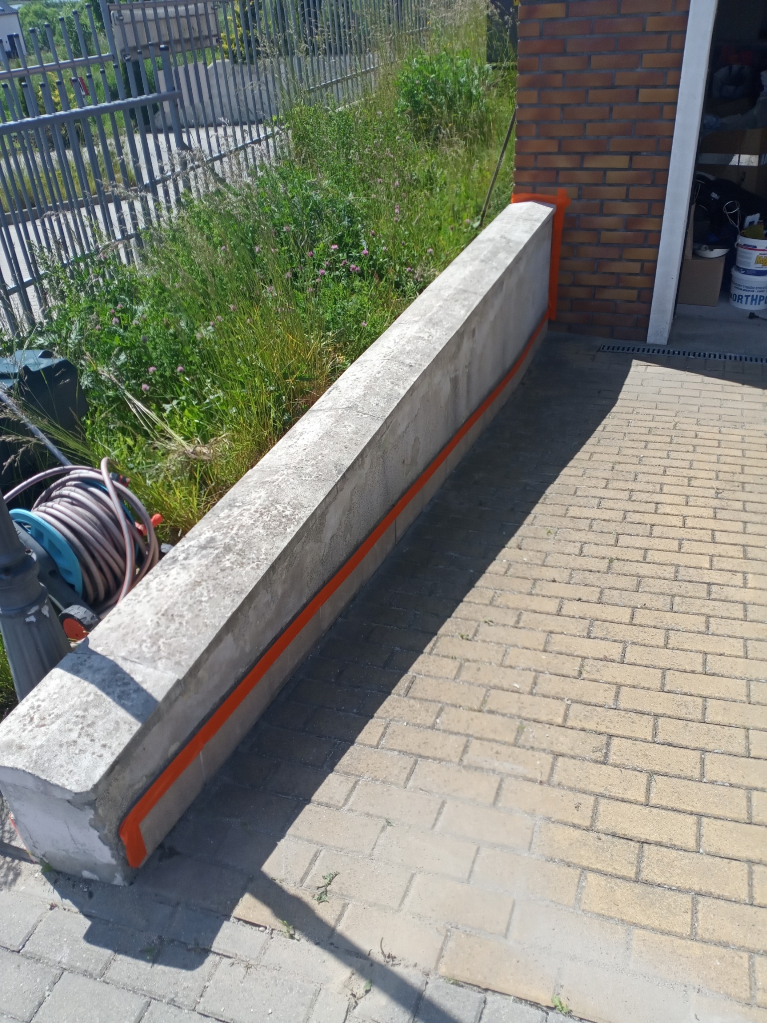 Betonowy murek z pomalowaną na pomarańczowo listwą, oddzielający posesję od chodnika z kostki. W tle widoczne trawy i metalowe ogrodzenie.
