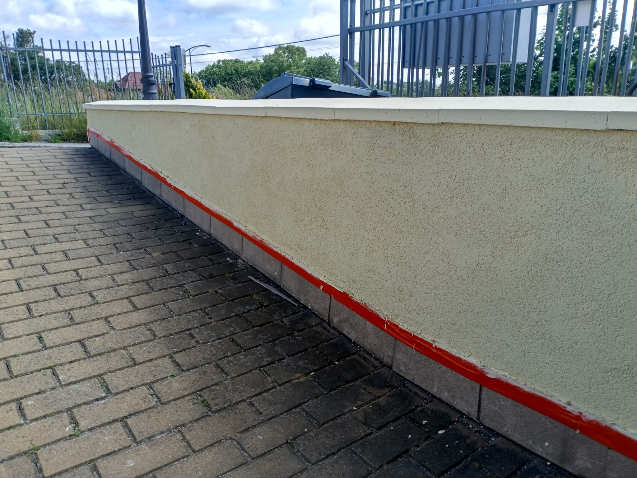 Otynkowany murek z czerwoną linią, widoczny z perspektywy chodnika z kostki brukowej, na tle metalowego ogrodzenia i zieleni.