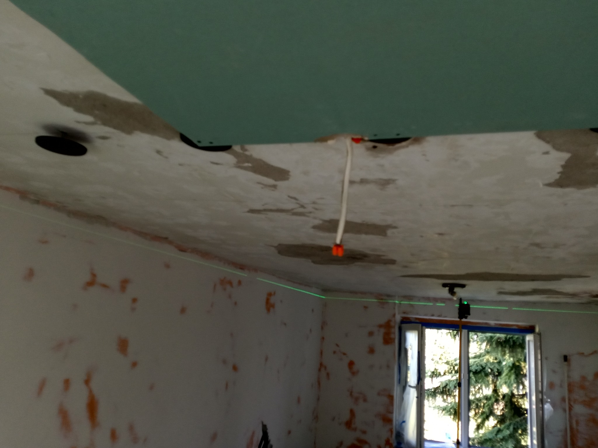 Montaż płyt gipsowych na suficie, widoczny laser krzyżowy i przewody elektryczne. Ściany przygotowane do malowania, okno z widokiem na drzewo.