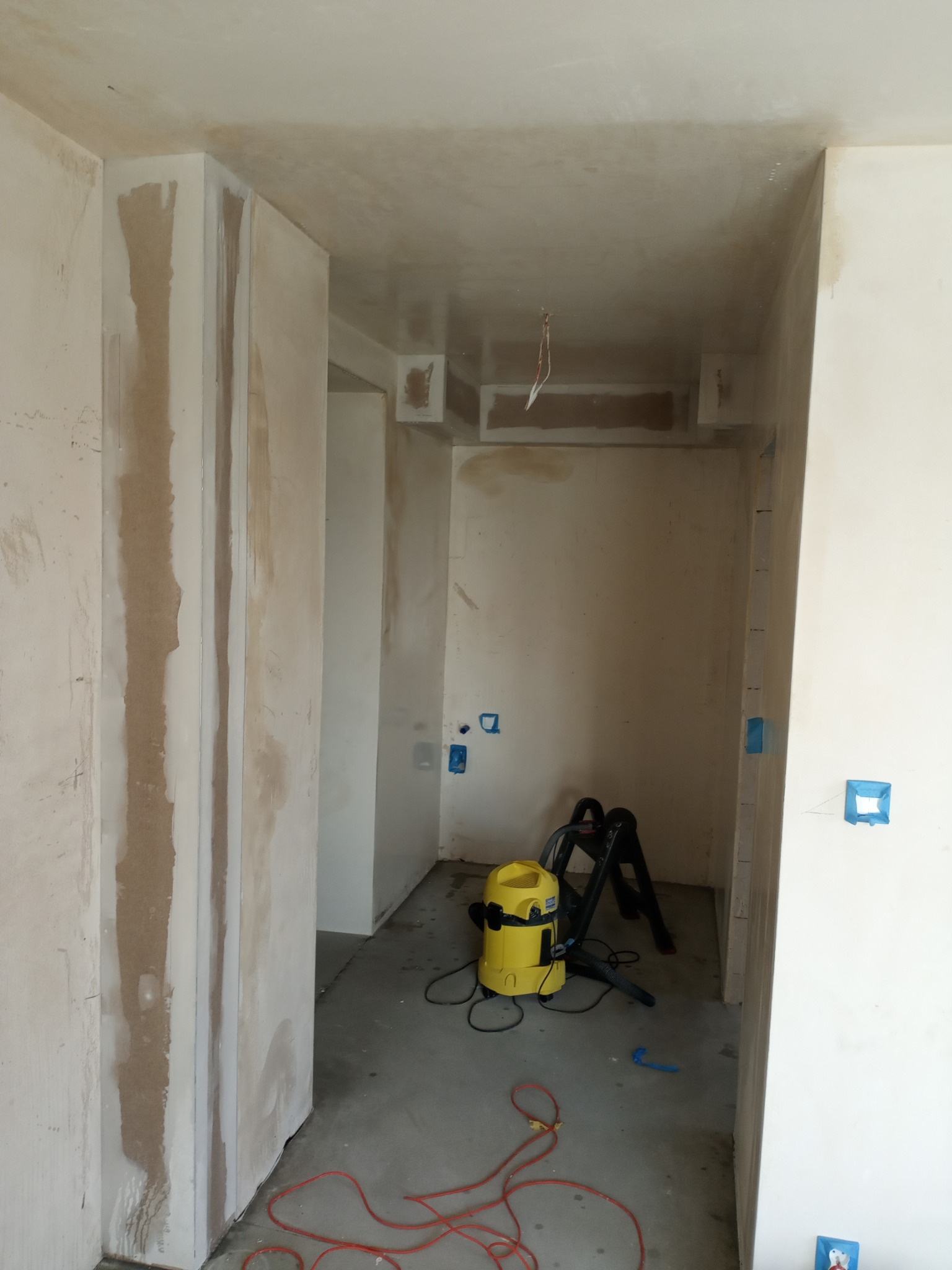 Wnętrze w trakcie prac wykończeniowych: ściany po szpachlowaniu, widoczny odkurzacz przemysłowy i drabina. Przygotowanie do malowania.