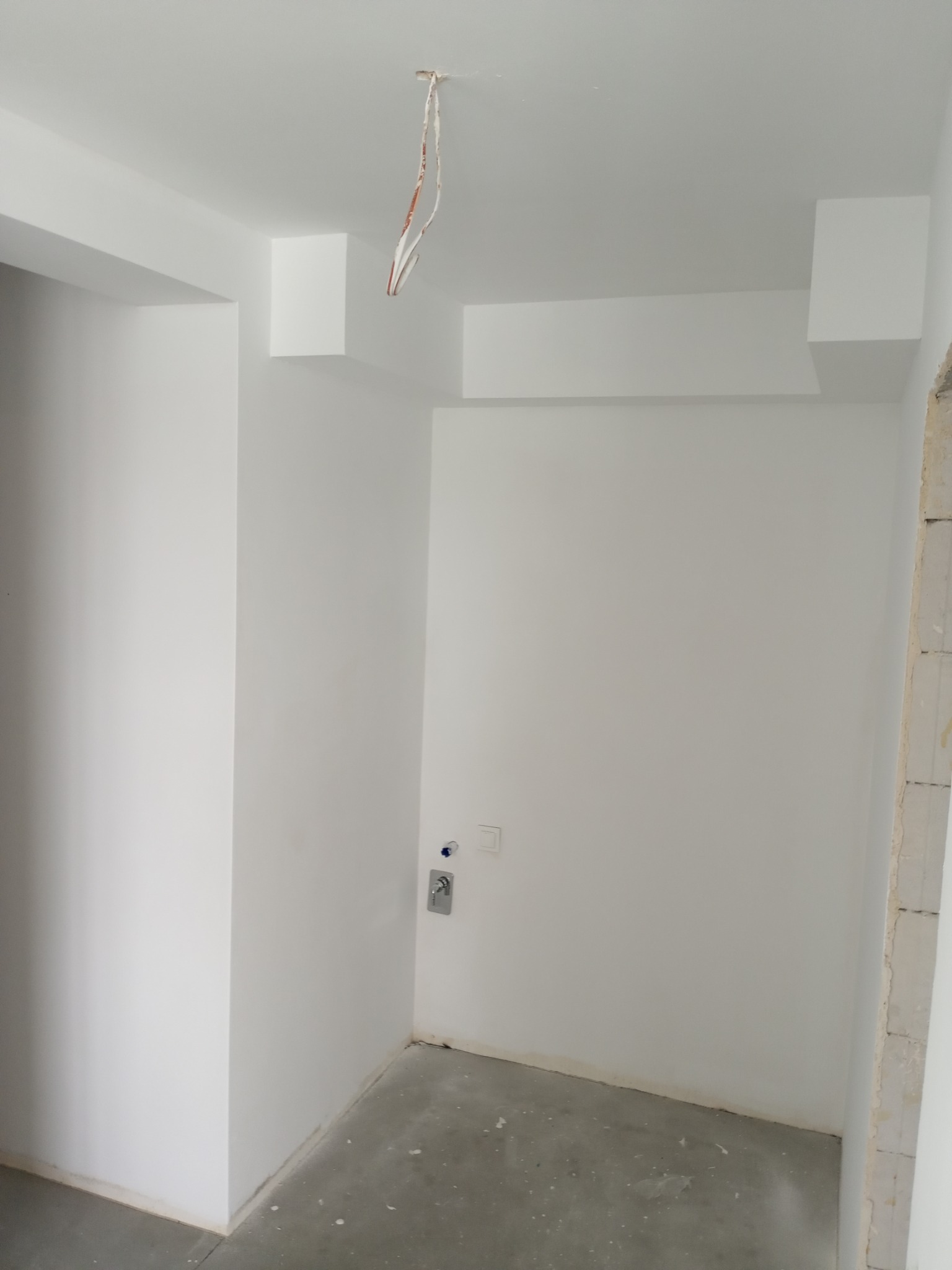 Białe wnętrze po malowaniu z wystającymi kablami z sufitu, widoczny fragment ściany z instalacją wodną i włącznikiem światła. Minimalistyczny design.