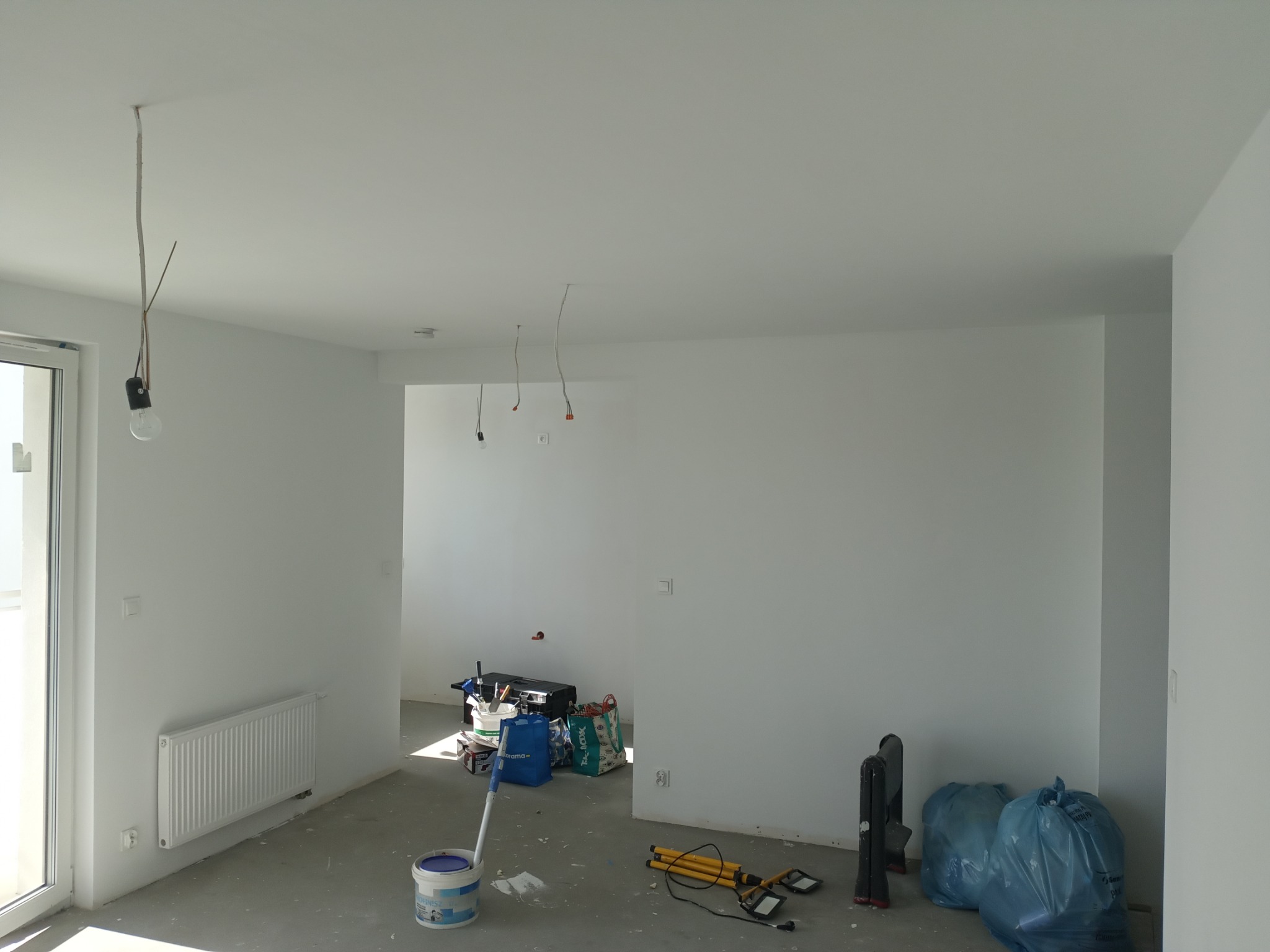 Wnętrze po malowaniu, widoczne białe ściany, grzejnik, okno balkonowe, narzędzia malarskie i worki na śmieci. Kabel z żarówką zwisa z sufitu.