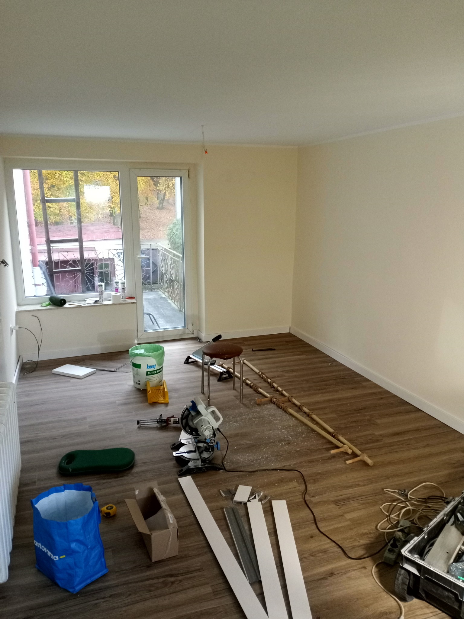Pokój w trakcie remontu: panele podłogowe, listwy przypodłogowe, narzędzia, wiadro z farbą. Widok na balkon i drzewa za oknem. Etap prac wykończeniowych.
