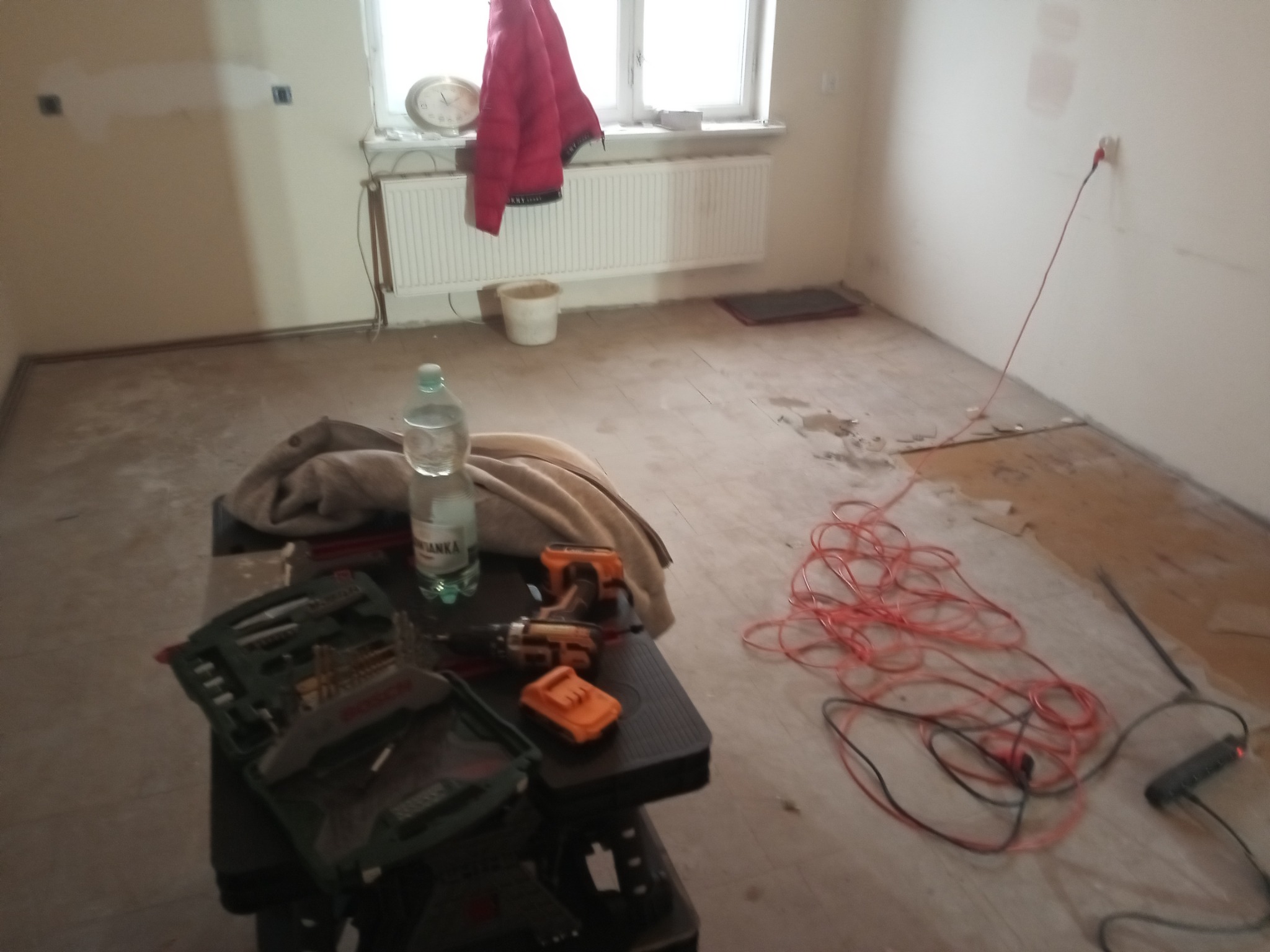 Pokój w trakcie remontu: odsłonięta podłoga, narzędzia (wiertarka, skrzynka z bitami) na stole, przedłużacz na podłodze. Widoczne ślady po demontażu podłogi.