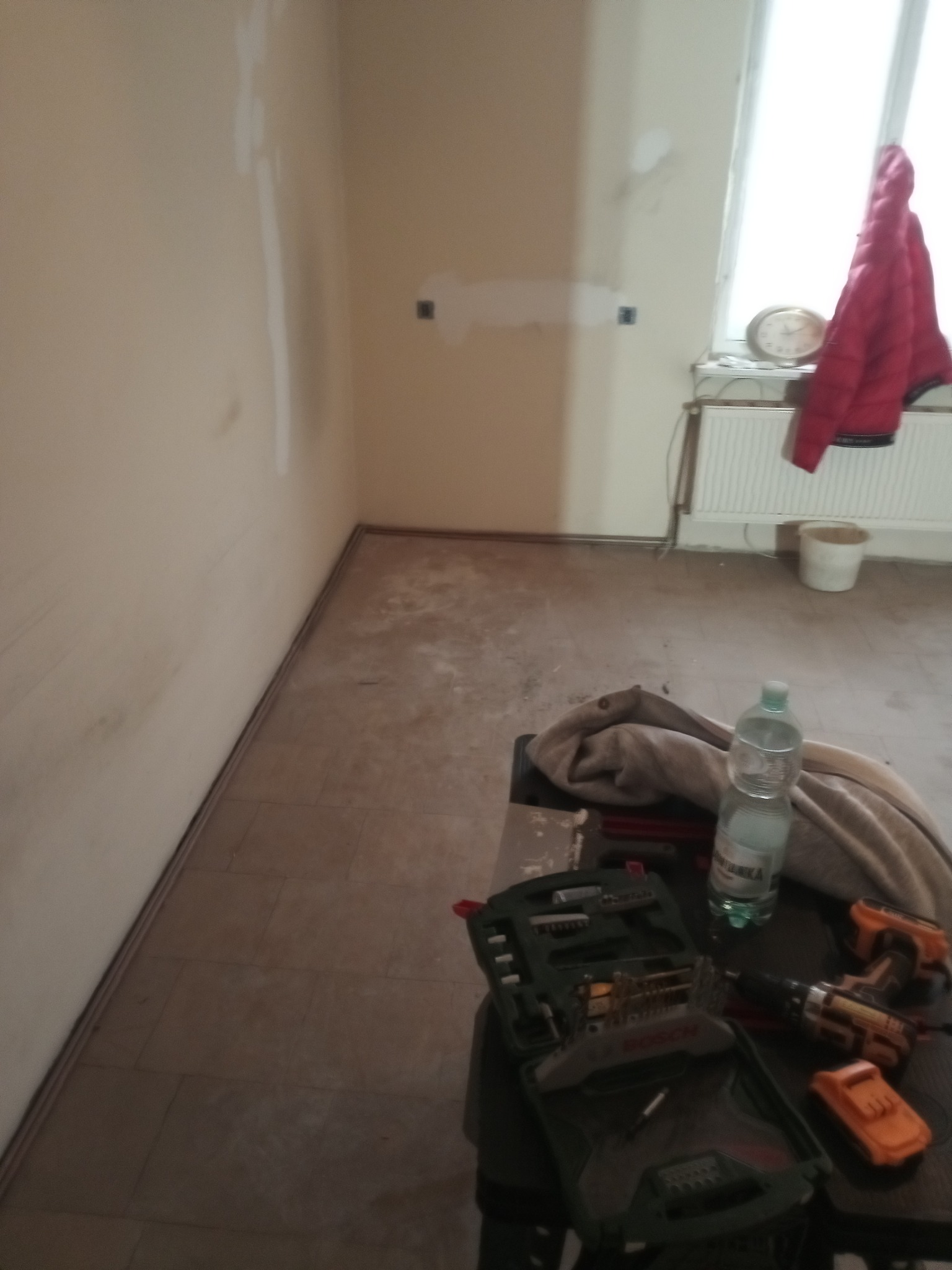 Pokój w trakcie remontu: przygotowane ściany, częściowo zdjęta podłoga, narzędzia na stole. Widoczne ślady po gładzi i przygotowania do dalszych prac wykończeniowych.