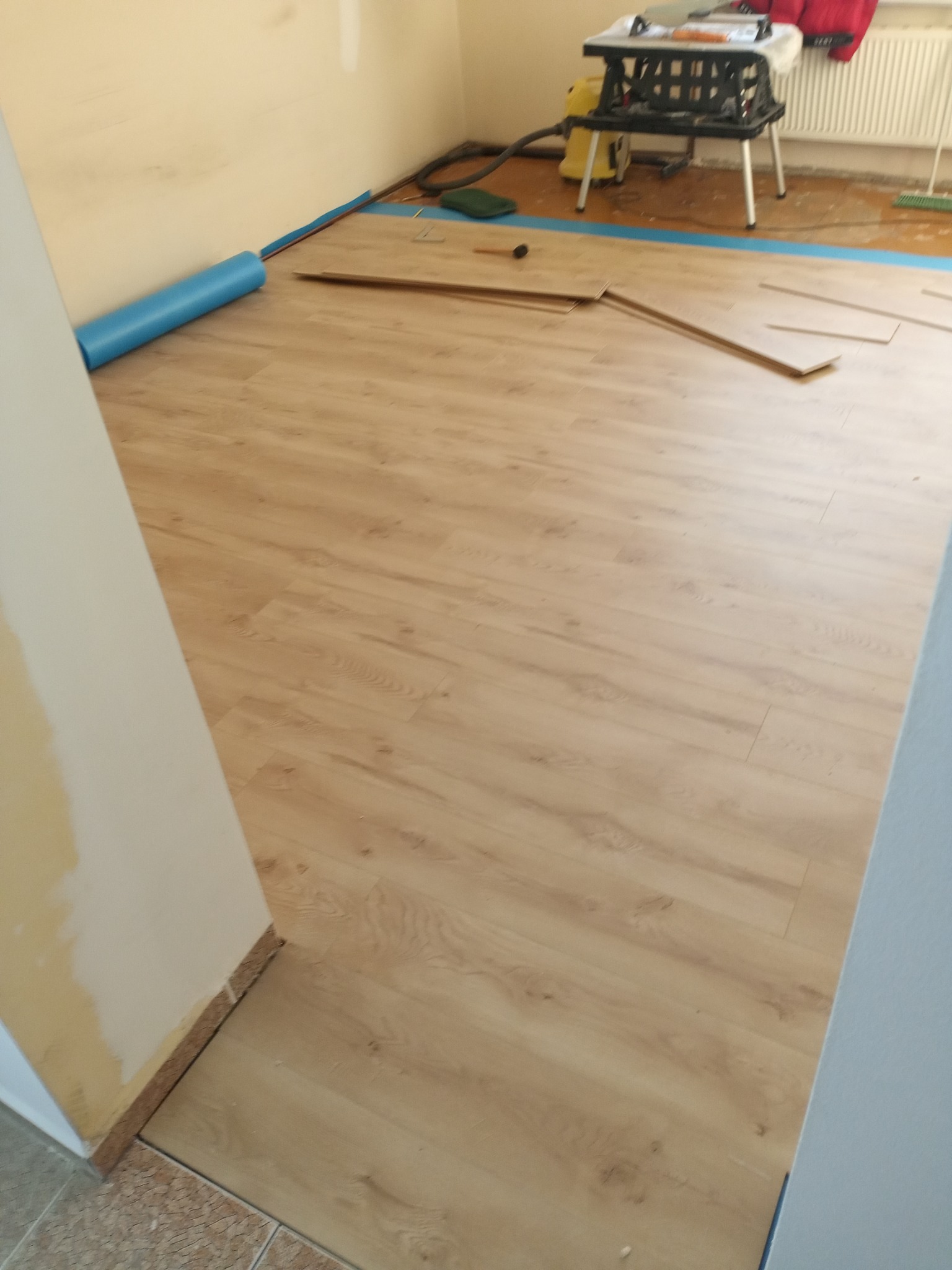 Układanie paneli podłogowych w trakcie prac. Widoczne panele, podkład, listwy przypodłogowe i narzędzia. Częściowo ułożona podłoga z paneli w jasnym odcieniu drewna.