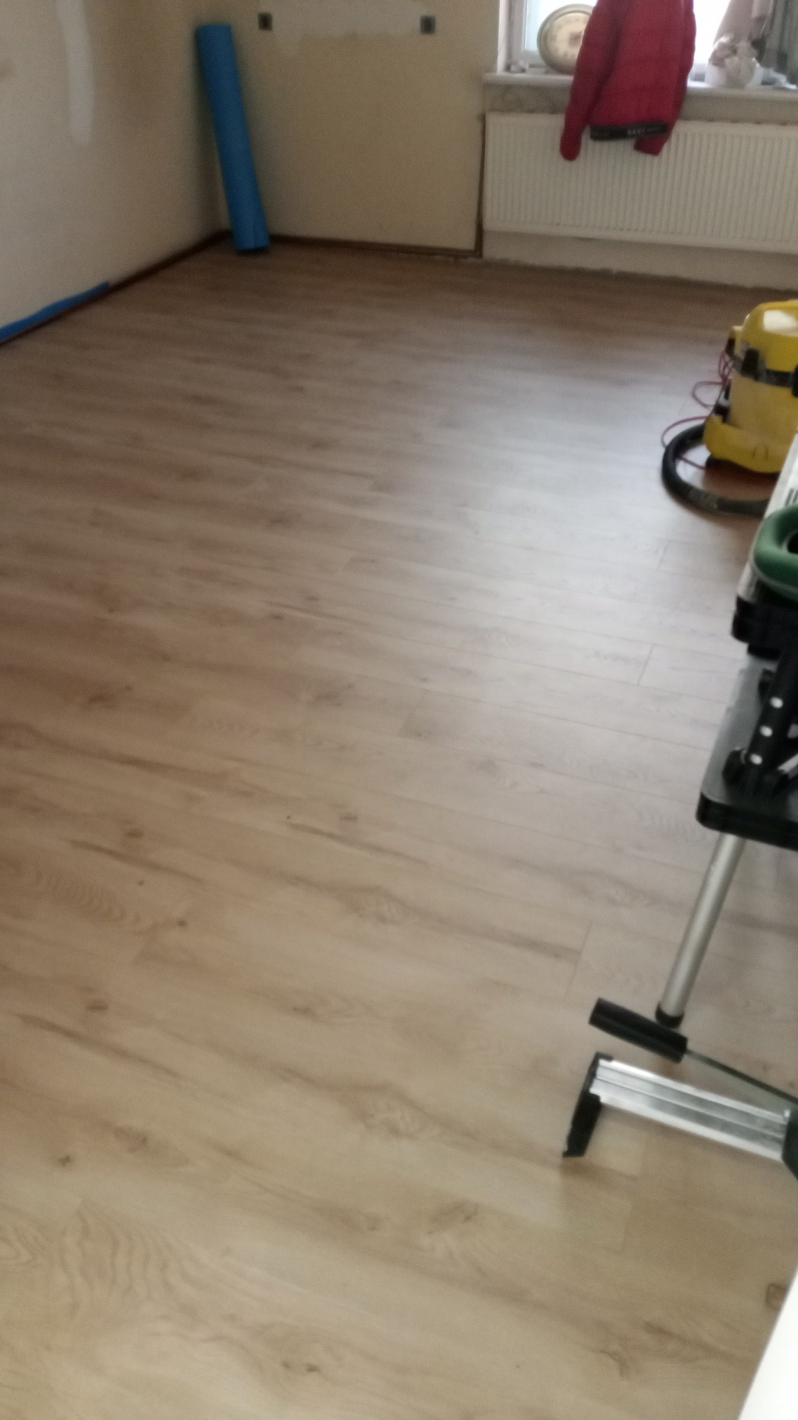 Jasne panele podłogowe w pokoju po remoncie, widoczny fragment drabiny malarskiej i odkurzacz. Ściany przygotowane do malowania, zabezpieczone folią.