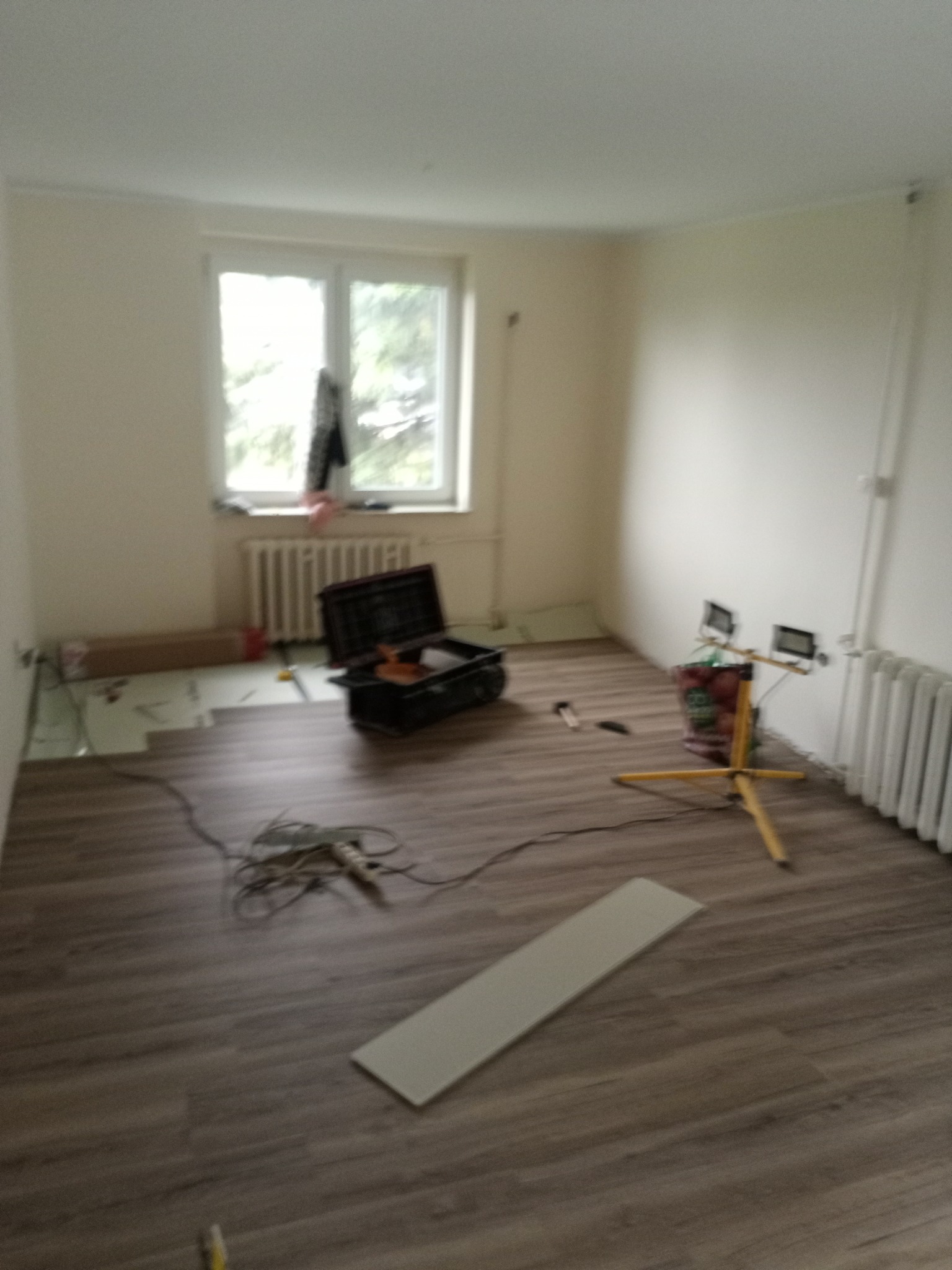 Układanie paneli podłogowych w pokoju. Częściowo ułożone panele w odcieniach brązu, narzędzia pracy, lampa oświetleniowa. Widok na okno i grzejnik.