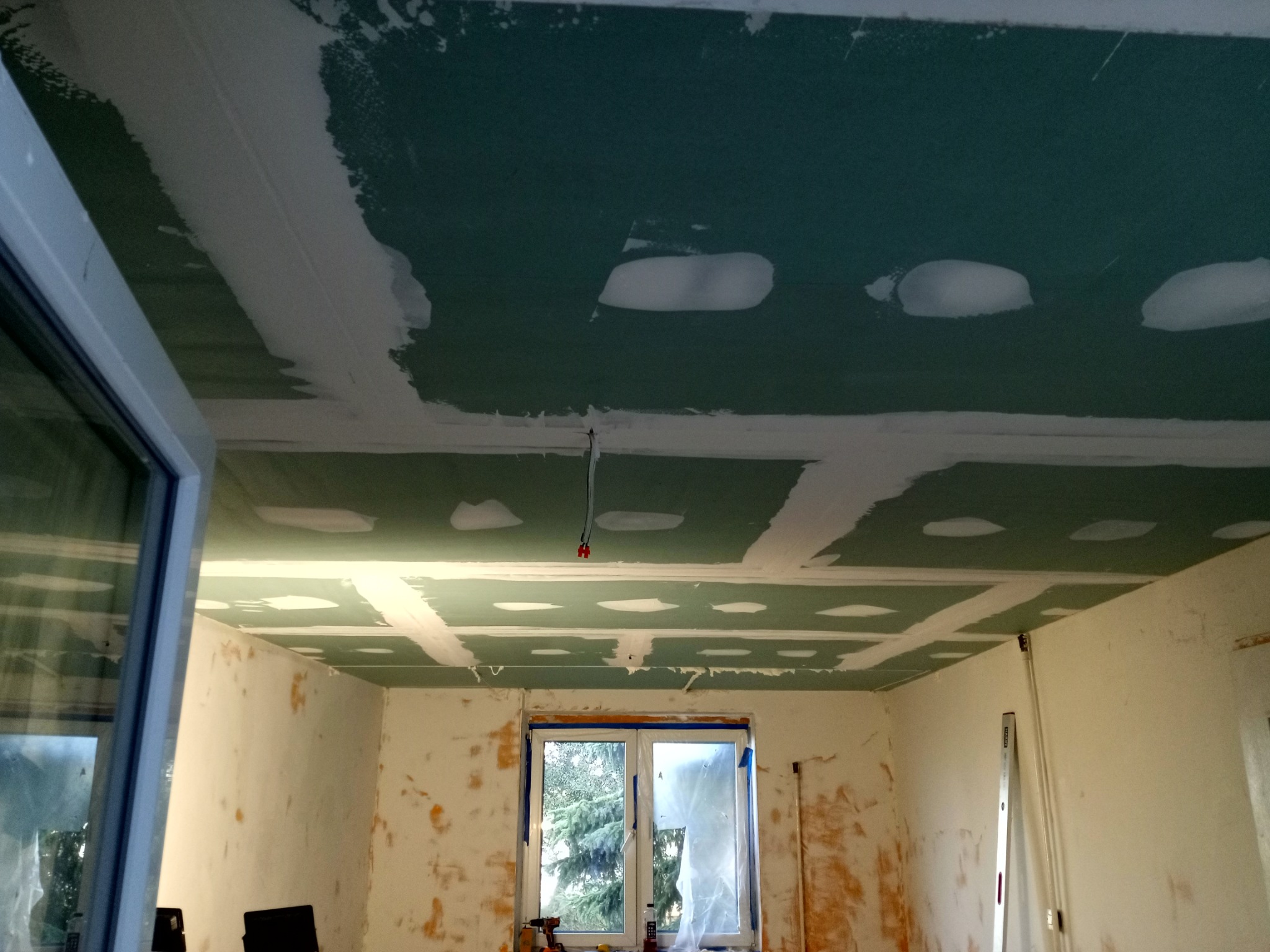 Montaż płyt karton-gips na suficie z widocznymi łączeniami i szpachlowaniem. W tle okno i ściany w trakcie przygotowania do dalszych prac wykończeniowych.