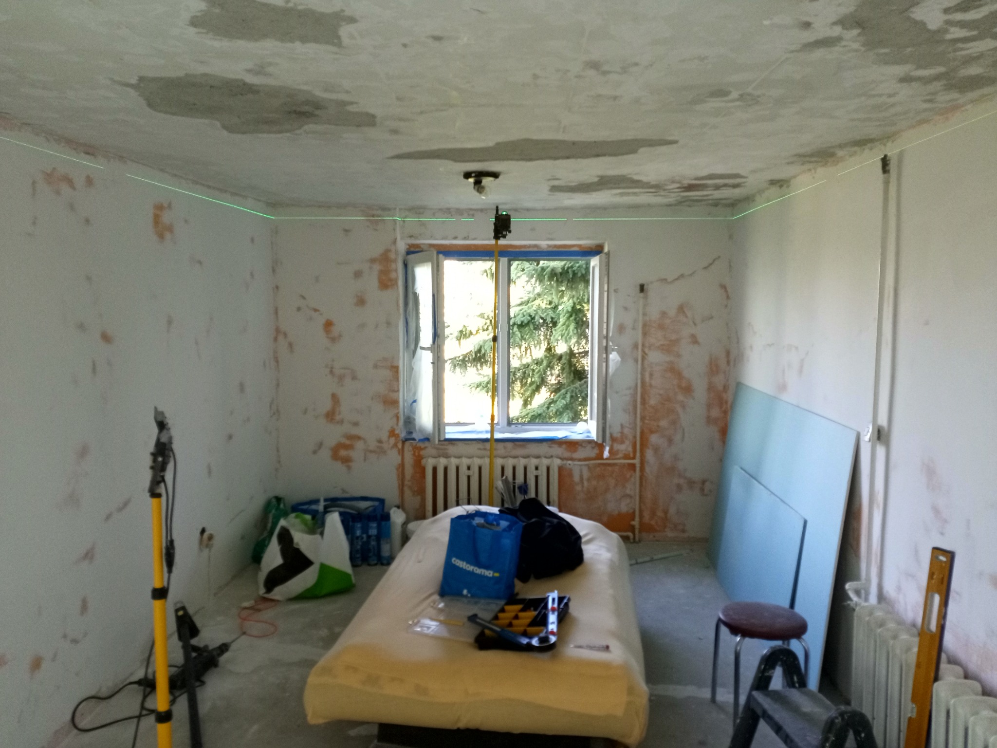 Pokój w trakcie remontu: ściany z resztkami farby, laserowy poziom, płyty karton-gips, narzędzia. Widoczne przygotowania do gładzi lub malowania.
