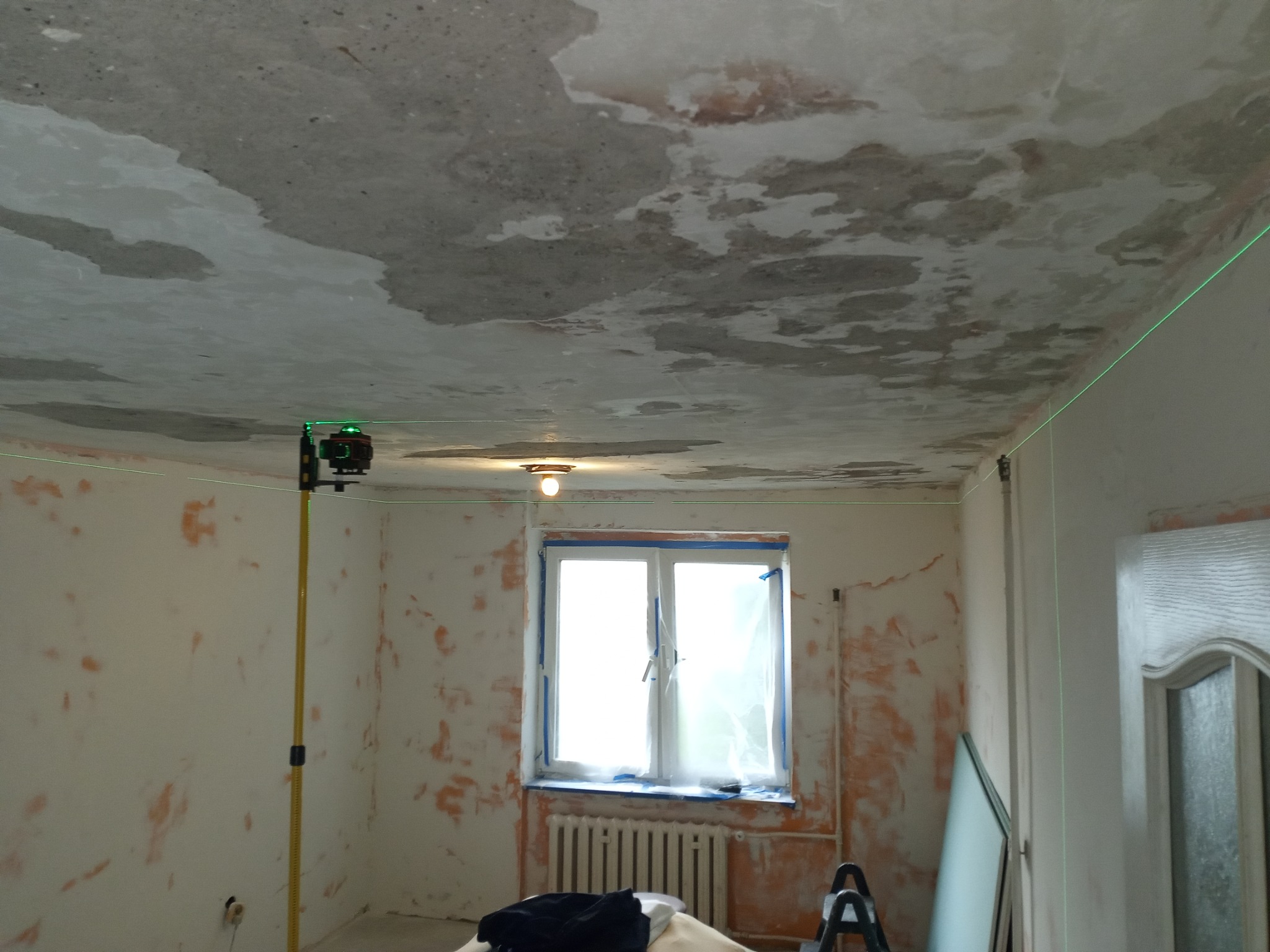 Pokój w trakcie remontu: ściany z resztkami farby, sufit z plamami, laser krzyżowy wyznacza poziom. Okno zabezpieczone folią malarską.