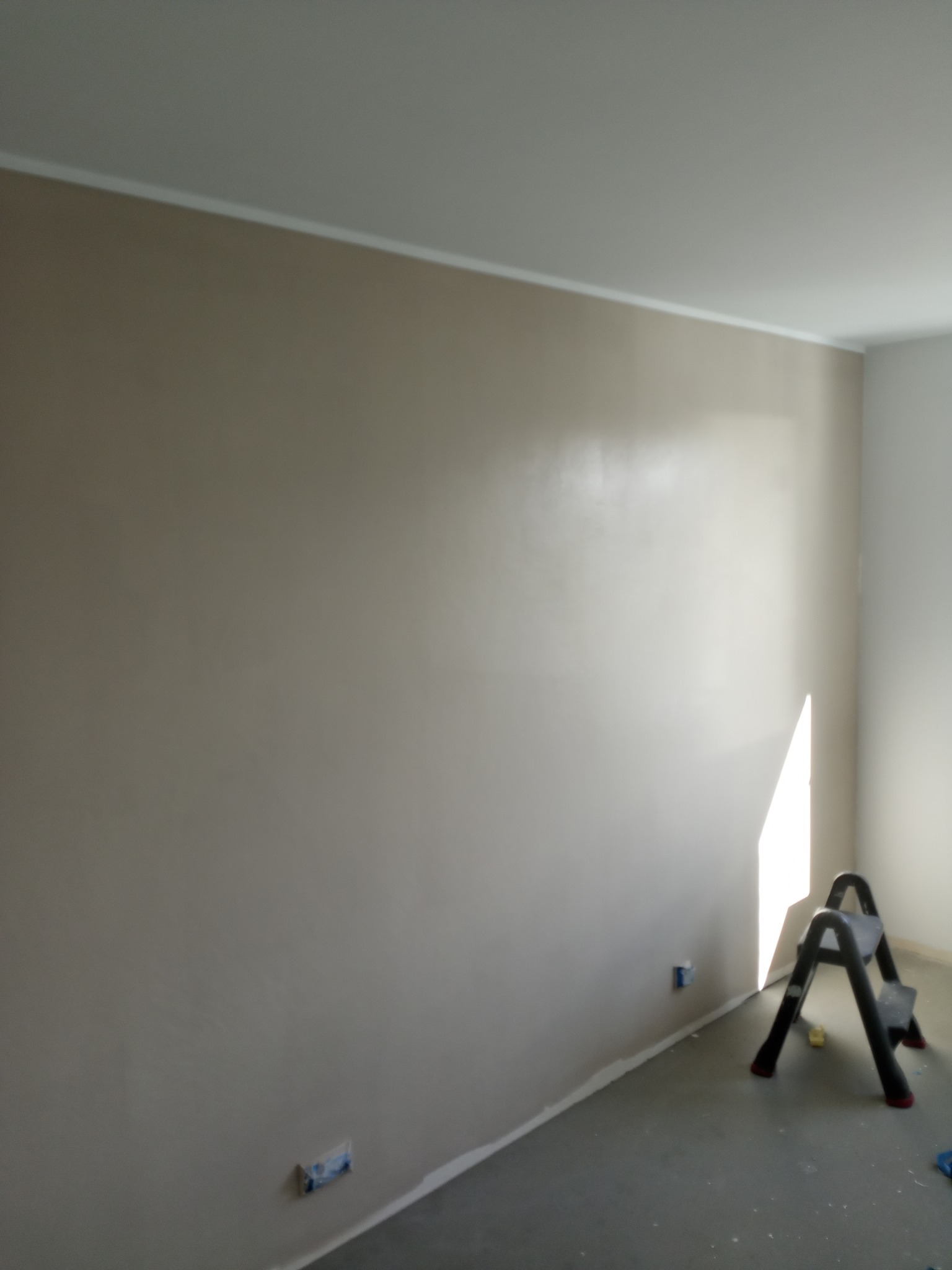 Ściana w odcieniach beżu po gładzi, widoczna drabina malarska i zabezpieczone gniazdka. Świeżo wykończone wnętrze z listwą przypodłogową.