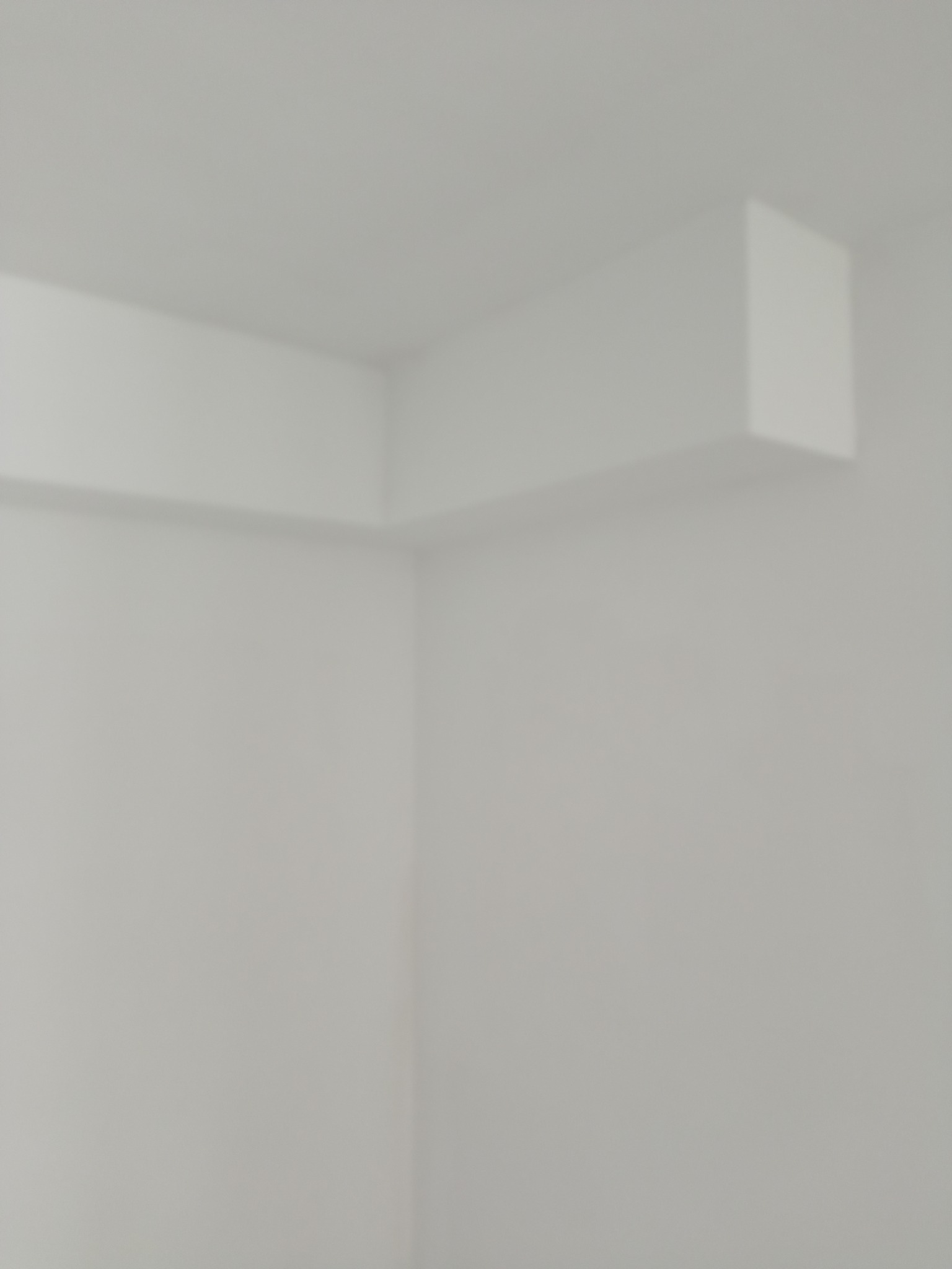 Minimalistyczne wnętrze: białe ściany i sufit z wystającym, pomalowanym na biało elementem konstrukcyjnym w rogu pomieszczenia. Delikatne światło.