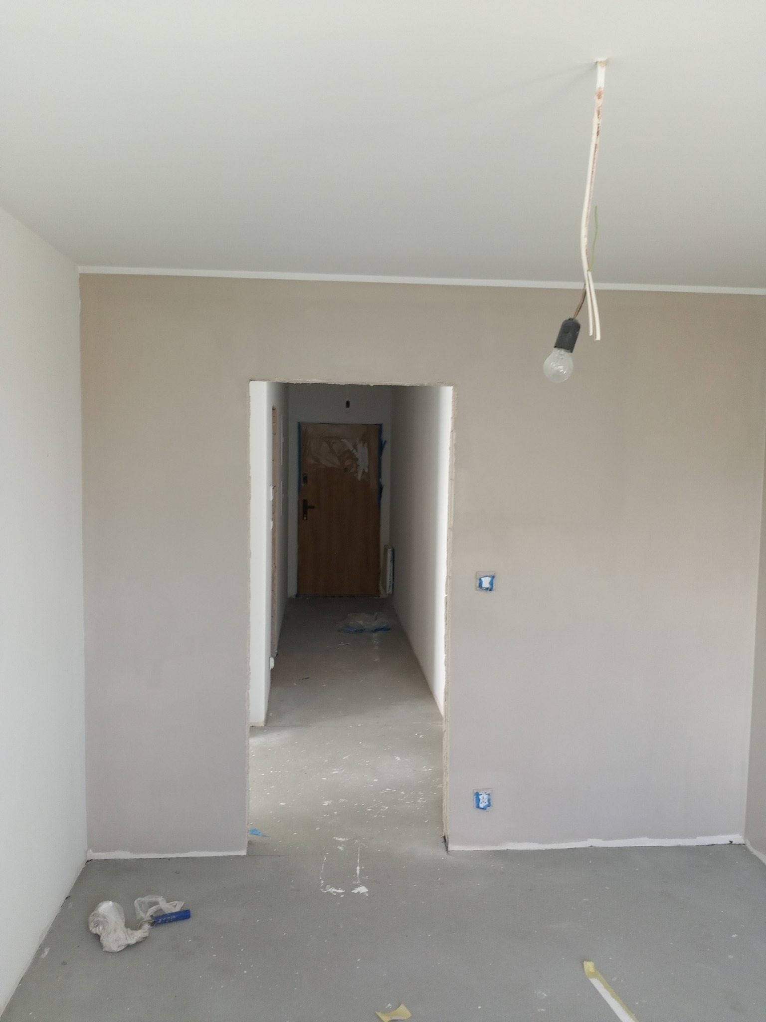Wnętrze po gładziowaniu ścian w odcieniach beżu i bieli, widoczne gniazdka elektryczne, surowa żarówka i ślady po pracy na podłodze. Widok na otwarte przejście do kolejnego pomieszczenia.