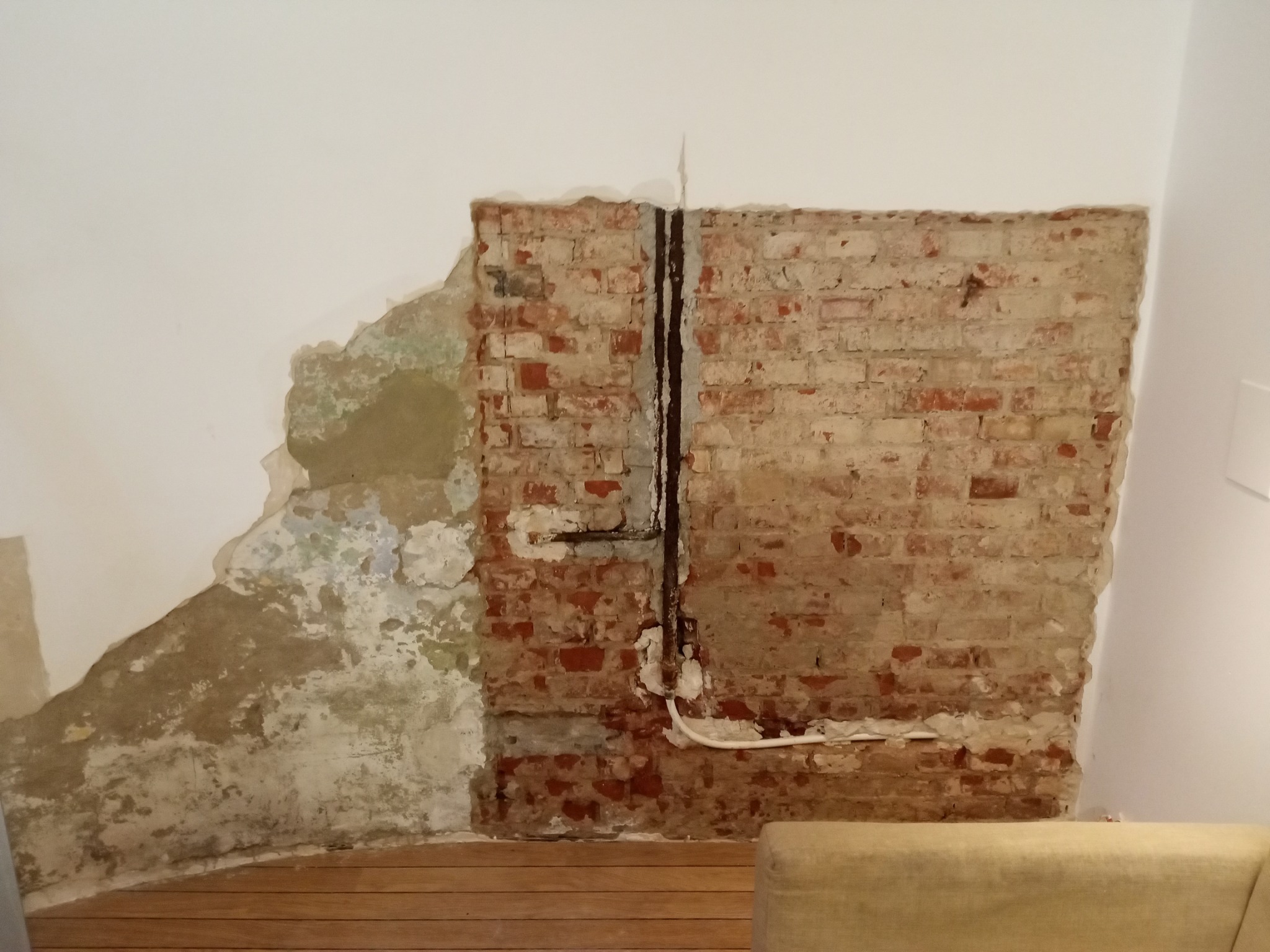 Odsłonięta cegła na ścianie z instalacją, fragmenty starego tynku i gładka, biała ściana. Widoczna drewniana podłoga i fragment mebla.