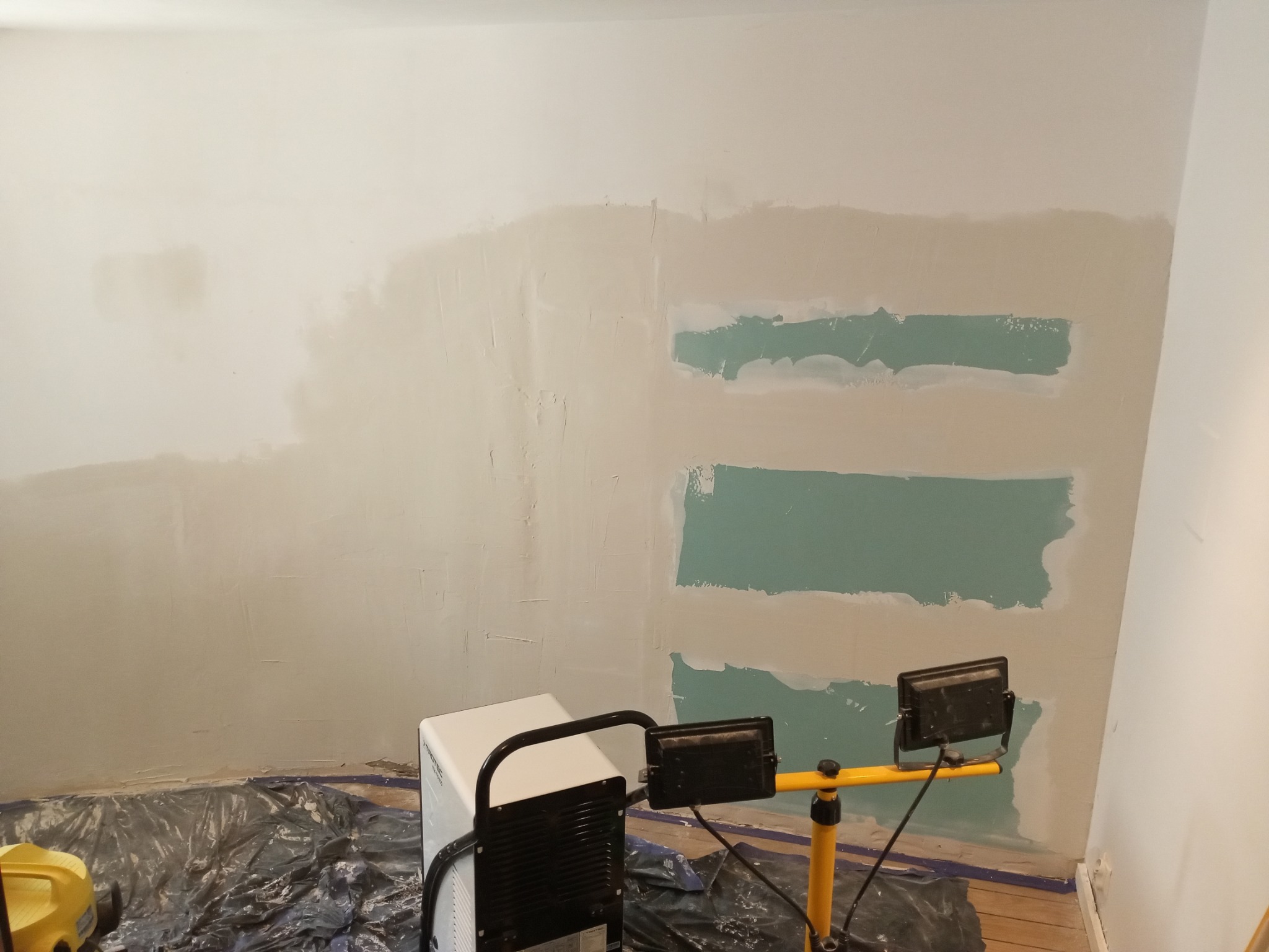 Ściana w trakcie szpachlowania z widocznymi fragmentami nałożonej gładzi, oświetlona lampą budowlaną. Podłoga zabezpieczona folią malarską.