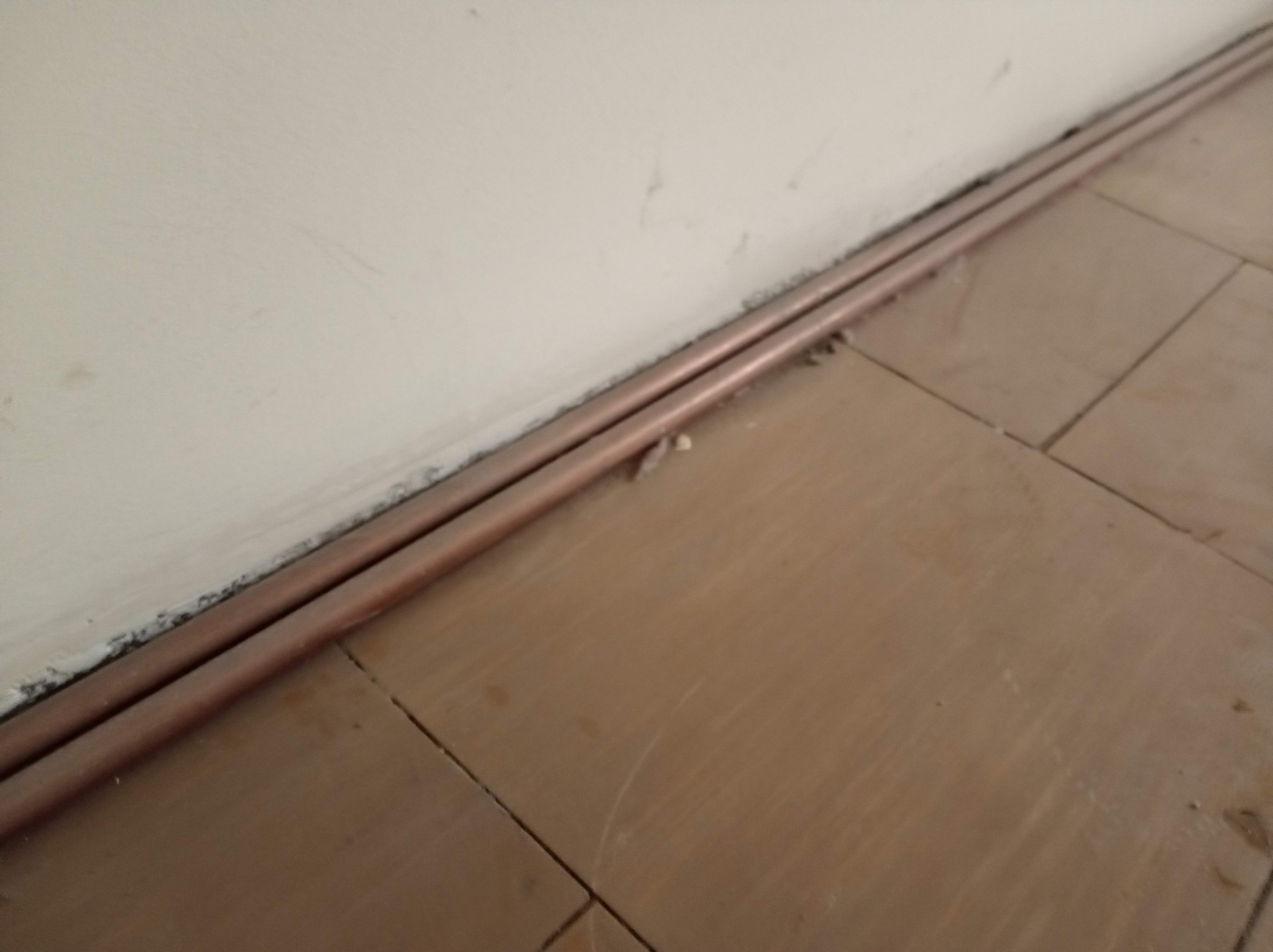 Miedziane rury grzewcze ułożone wzdłuż ściany na brązowych płytkach podłogowych. Widoczne ślady po remoncie przy ścianie.