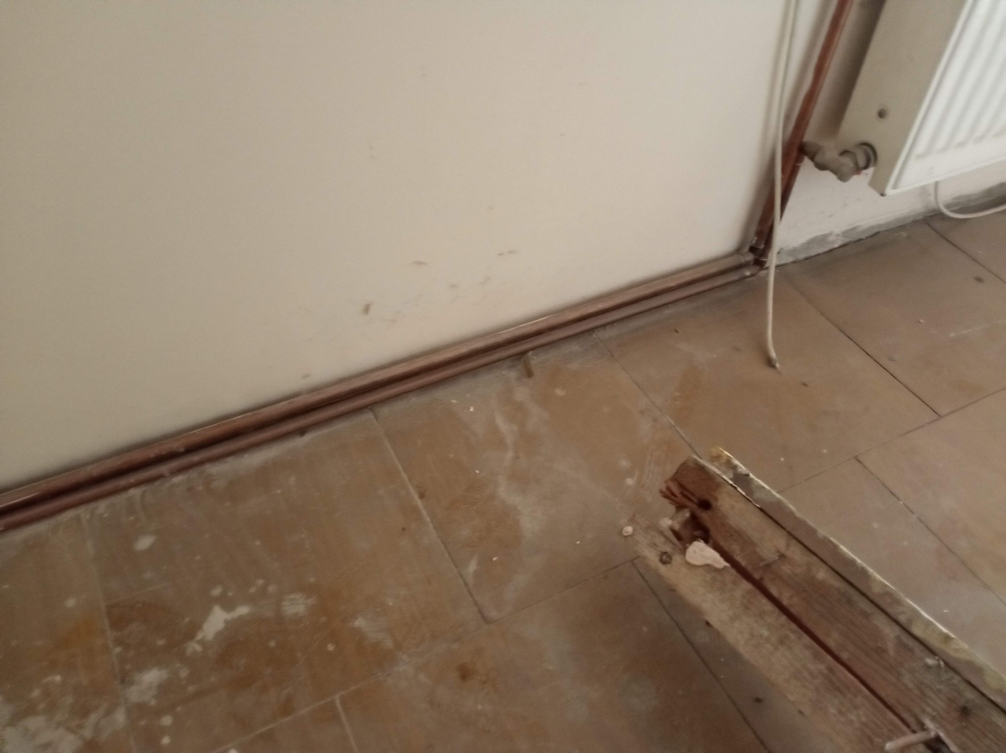 Fragment podłogi z płytkami, rurami grzewczymi przy ścianie i listwą przypodłogową w trakcie demontażu. Widoczne ślady po remoncie.