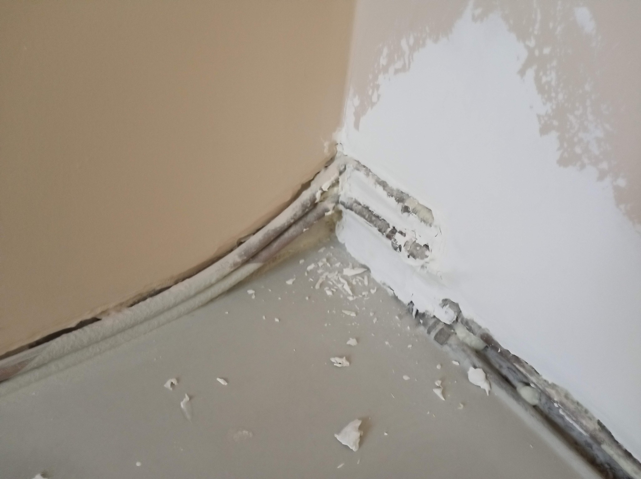 Narożnik ścian w trakcie prac wykończeniowych. Widoczne ślady po szpachlowaniu i malowaniu, zabezpieczone listwy przypodłogowe. Fragment podłogi z resztkami materiałów.