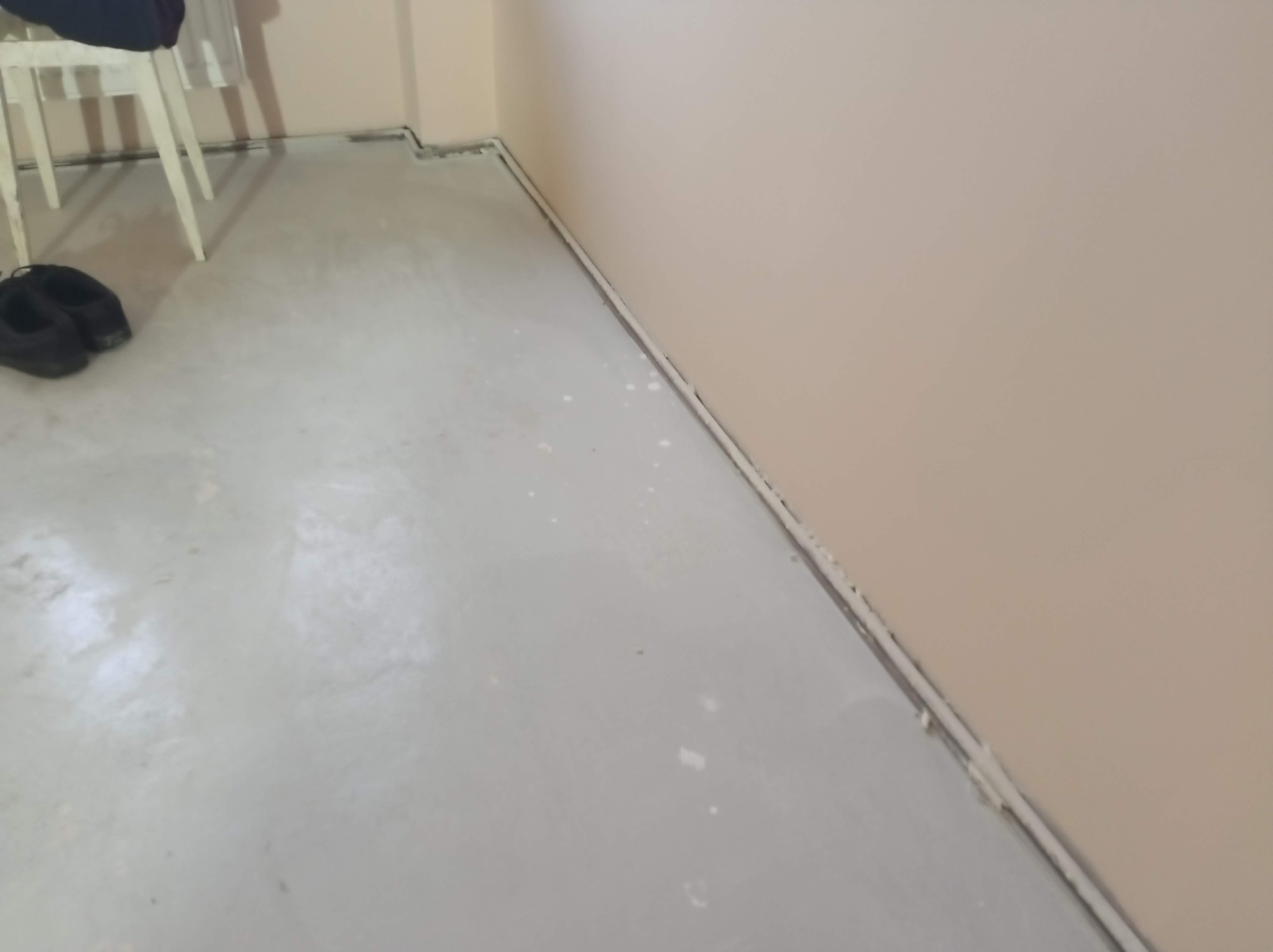Wylana, jasnoszara posadzka samopoziomująca w rogu pomieszczenia z beżową ścianą. Widoczne listwy przypodłogowe i buty robocze.