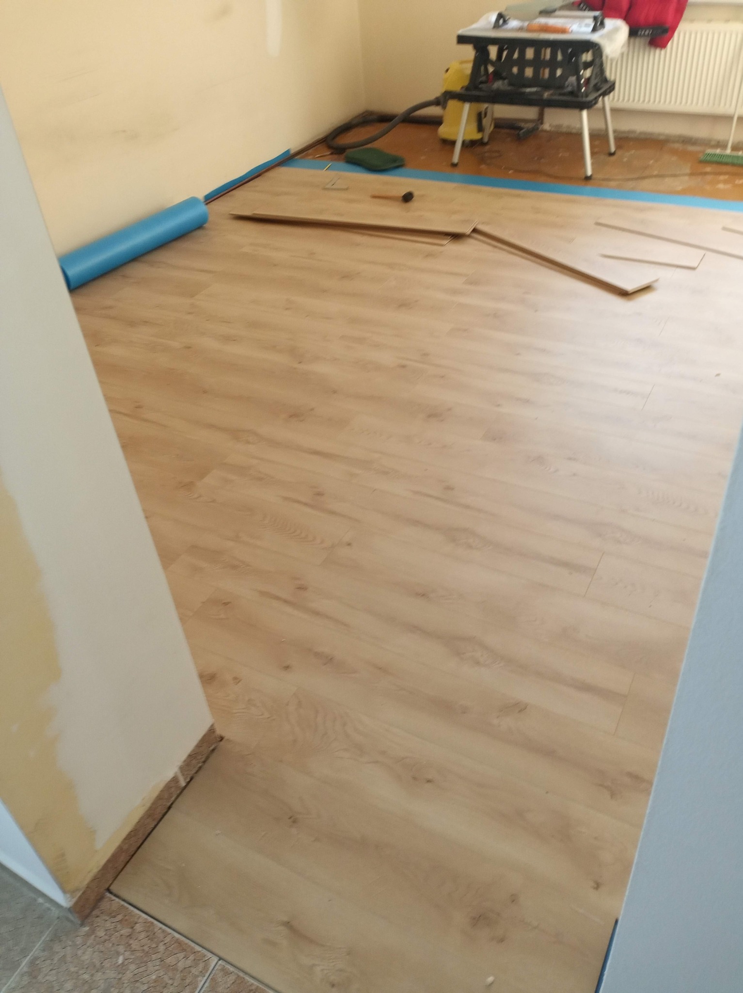 Układanie paneli podłogowych w jasnym odcieniu drewna, widoczne narzędzia i materiały przygotowane do montażu, niebieska folia izolacyjna przy ścianach.