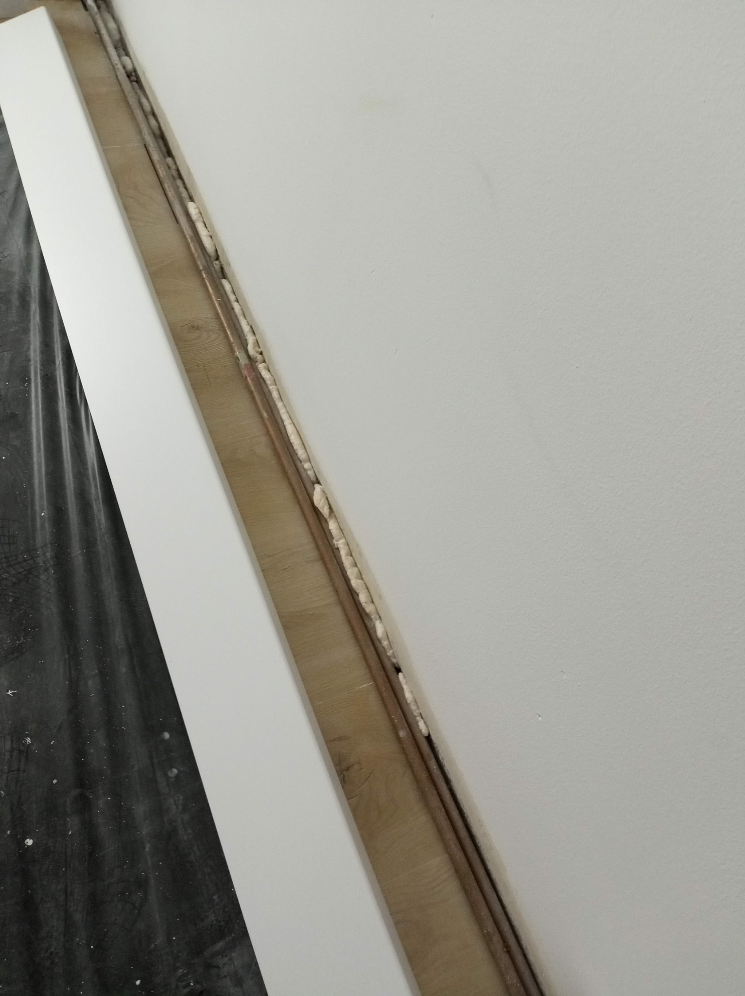 Biała ściana po gładziowaniu, z widocznymi rurami i pianką montażową w szczelinie. Fragment folii ochronnej w lewym dolnym rogu. Ujęcie z bliska, ukośne.