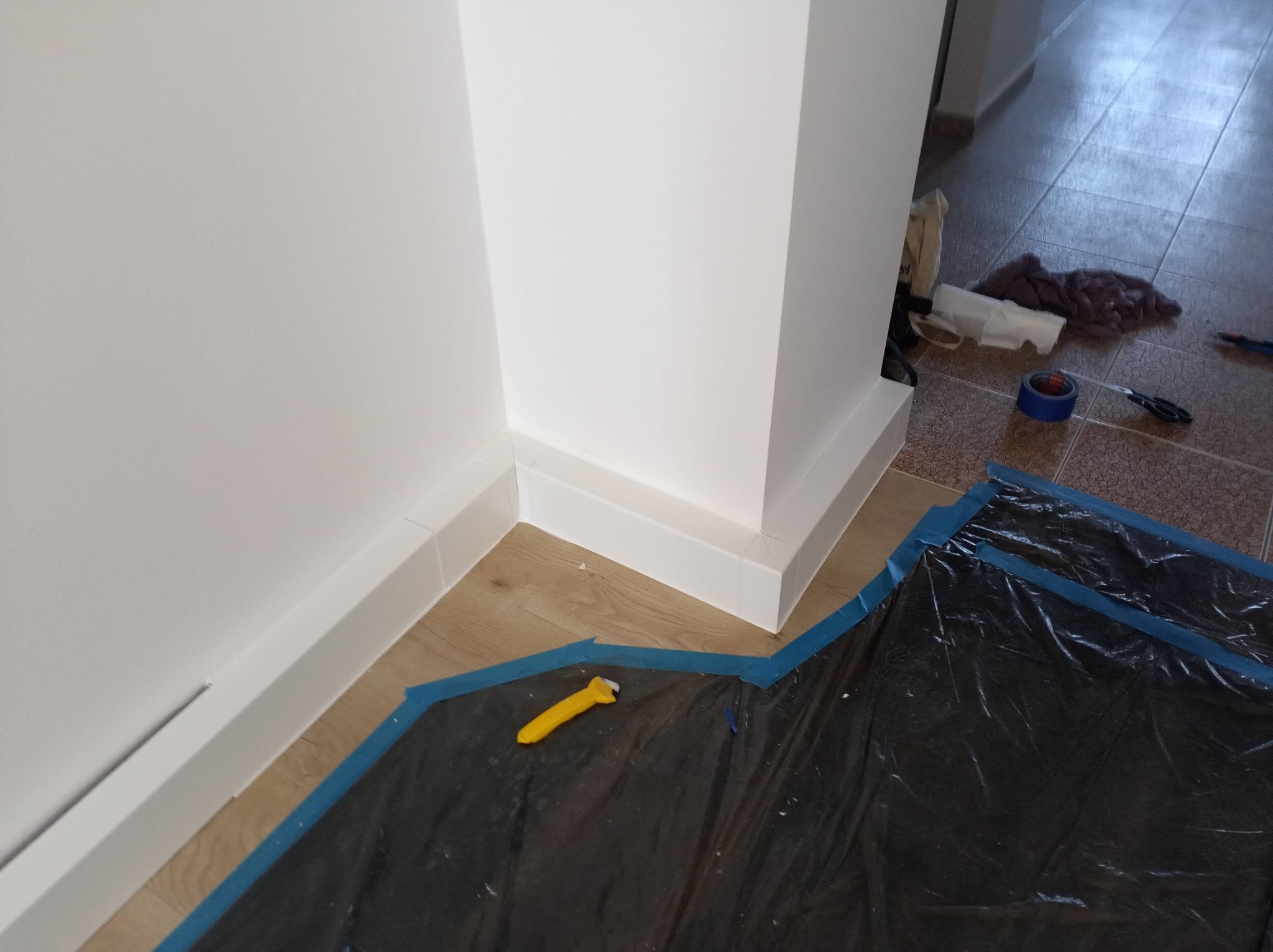 Białe listwy przypodłogowe i narożnik po malowaniu, zabezpieczone folią malarską. Widoczny nóż do tapet. Podłoga drewniana, w tle płytki.
