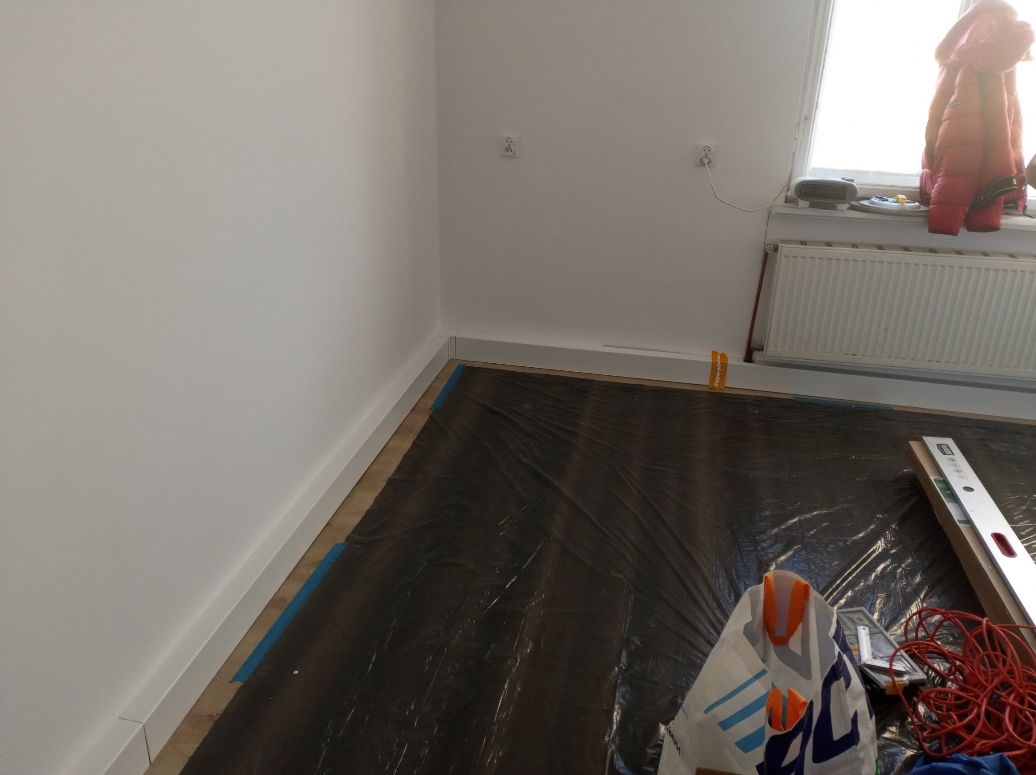 Pokój w trakcie prac wykończeniowych: zabezpieczona podłoga folią, listwy przypodłogowe zamontowane, widoczne narzędzia i poziomica. Ściany pomalowane na biało.