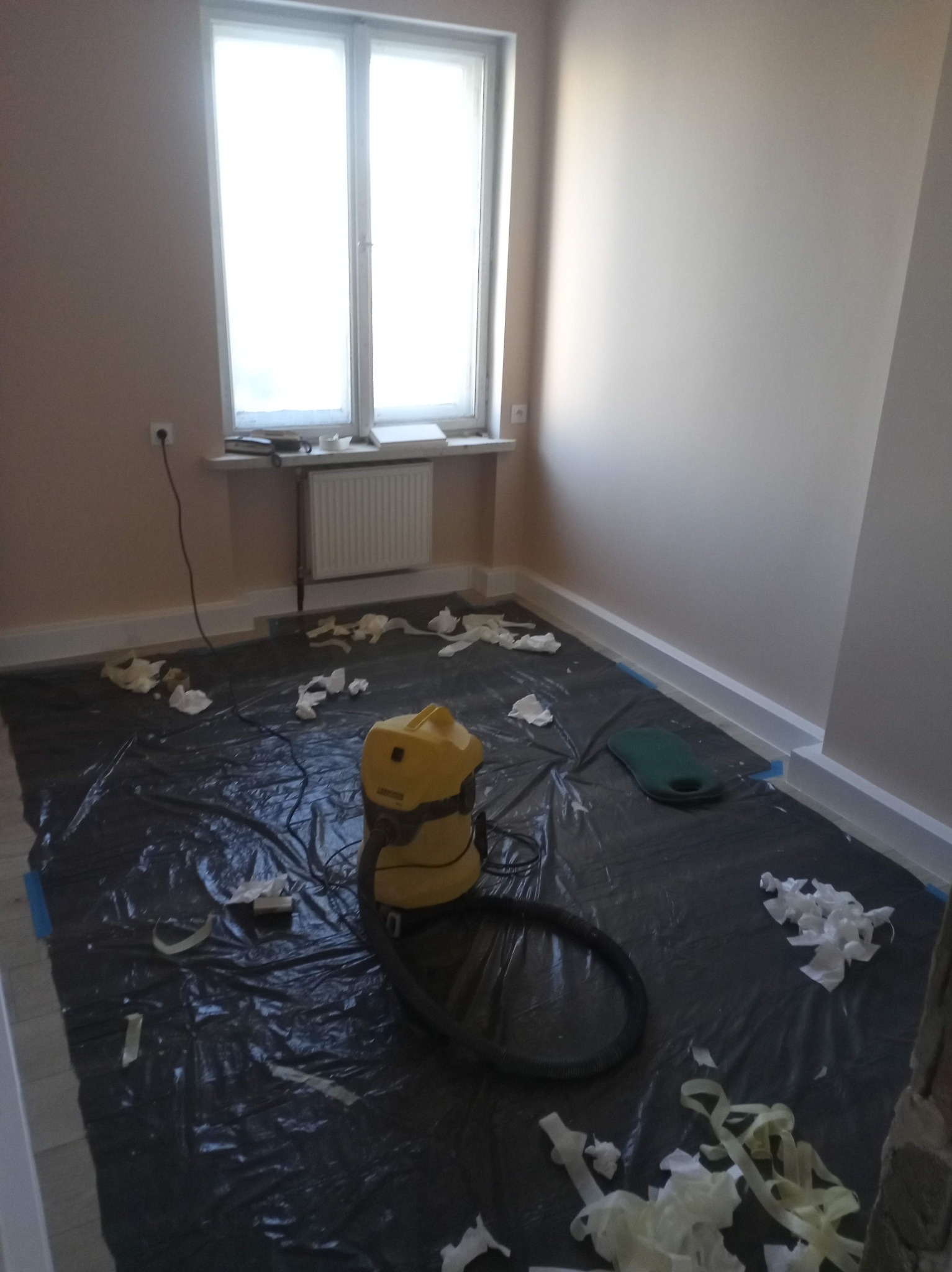 Pokój po malowaniu: zabezpieczona folią podłoga, odkurzacz przemysłowy, świeżo pomalowane ściany w odcieniach beżu, białe listwy przypodłogowe i okno.