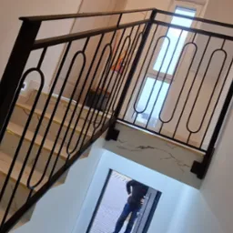 Czarna, metalowa balustrada schodowa z owalnymi zdobieniami, zamontowana na marmurowych schodach z podświetleniem LED w ścianie.