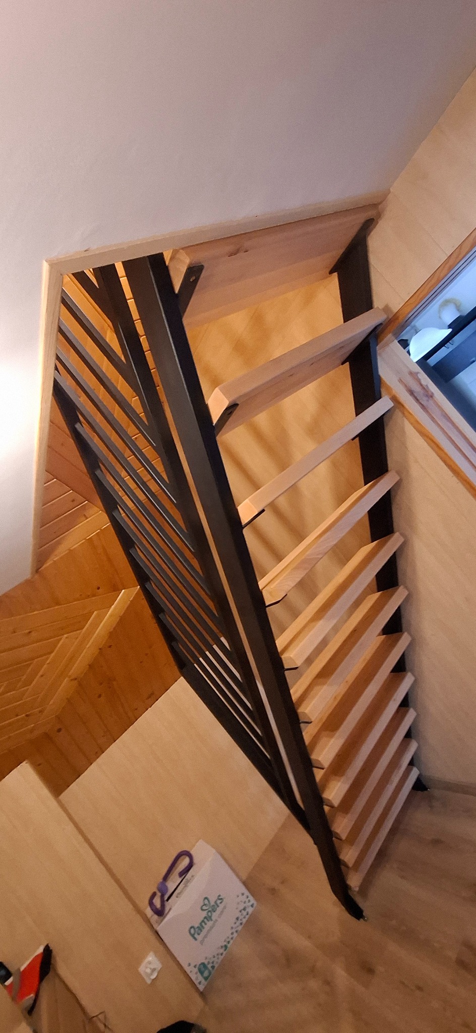 Ażurowe schody drewniane z czarną metalową balustradą, widok z dołu, częściowo widoczna paczka pieluch na podłodze.