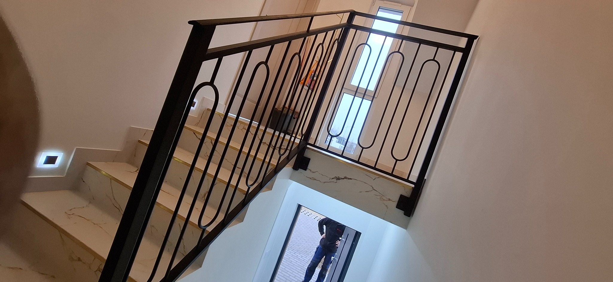 Czarna, metalowa balustrada schodowa z owalnymi zdobieniami, zamontowana na marmurowych schodach z podświetleniem LED w ścianie.