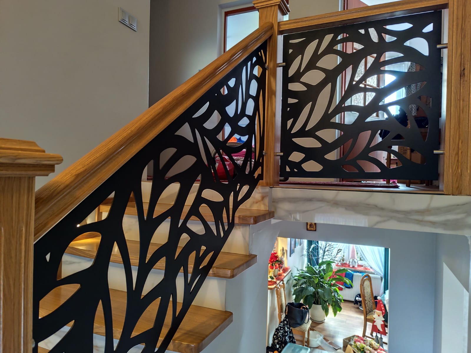 Nowoczesna balustrada schodowa z drewnianymi elementami i czarnym, ażurowym wypełnieniem o motywie liściastym, widok z dołu schodów.